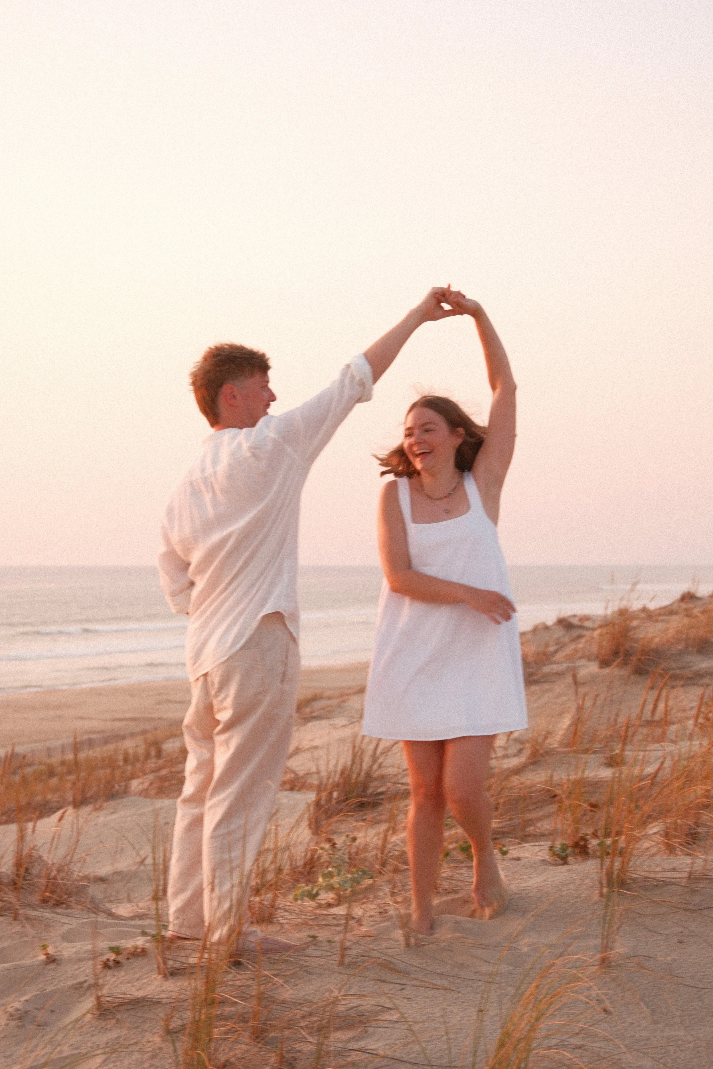 Un homme et une femme dansant sur la plage au coucher du soleil, souriant et tenus par la main, vêtus de vêtements légers blancs.