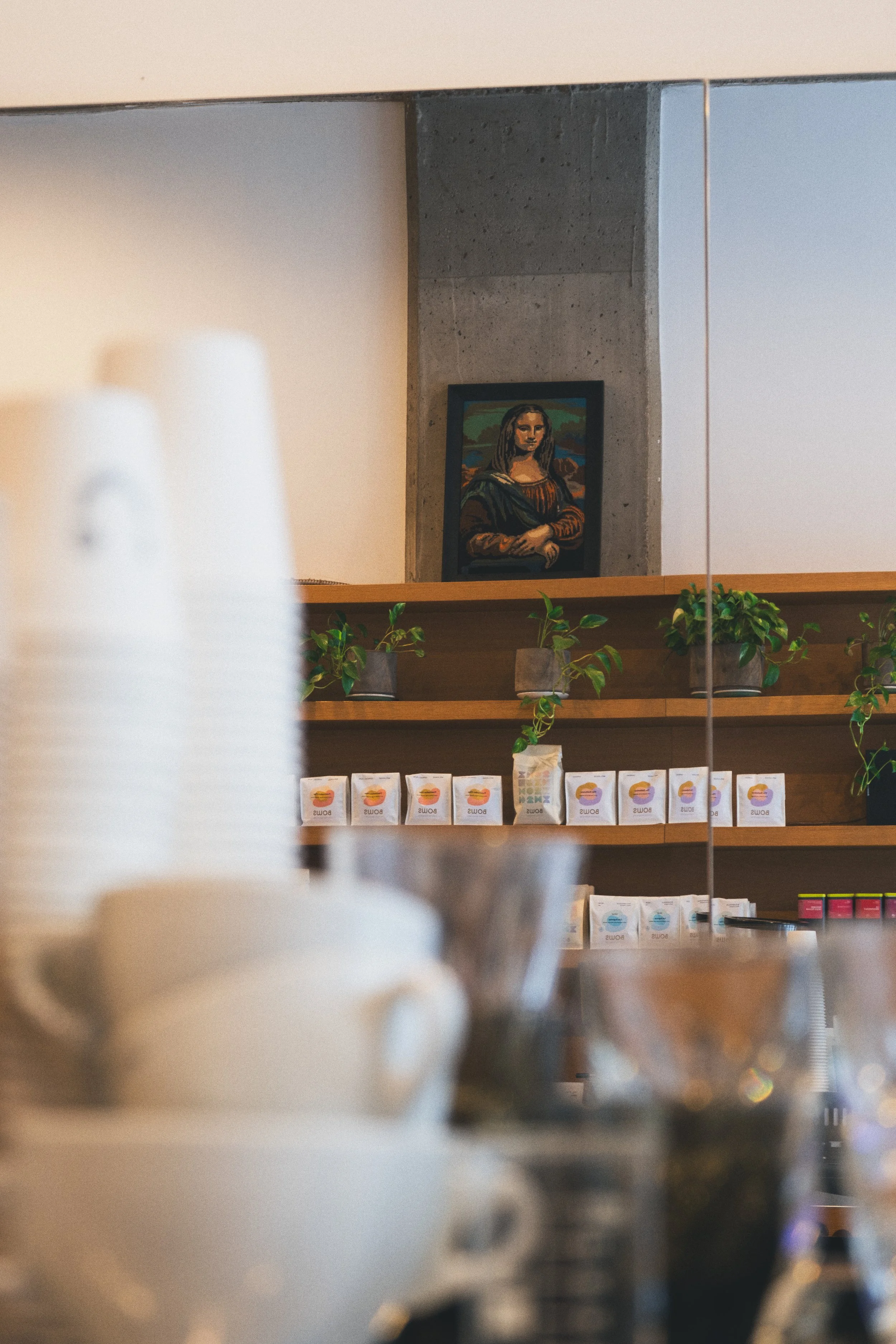 Une étagère en bois avec plusieurs boîtes de thé ou de café, décorée avec des plantes vertes. En arrière-plan, un mur en béton avec un tableau de la Joconde.
