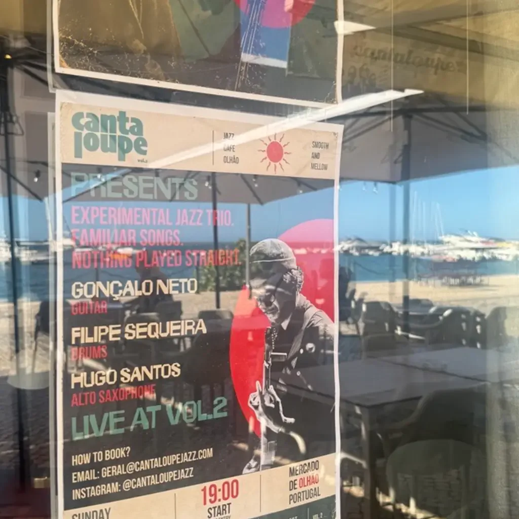 A poster behind a glass window advertising a live jazz performance at Mercado de Olhão in Portugal. The poster features a black and white image of a jazz musician playing guitar, with colorful text detailing the event.