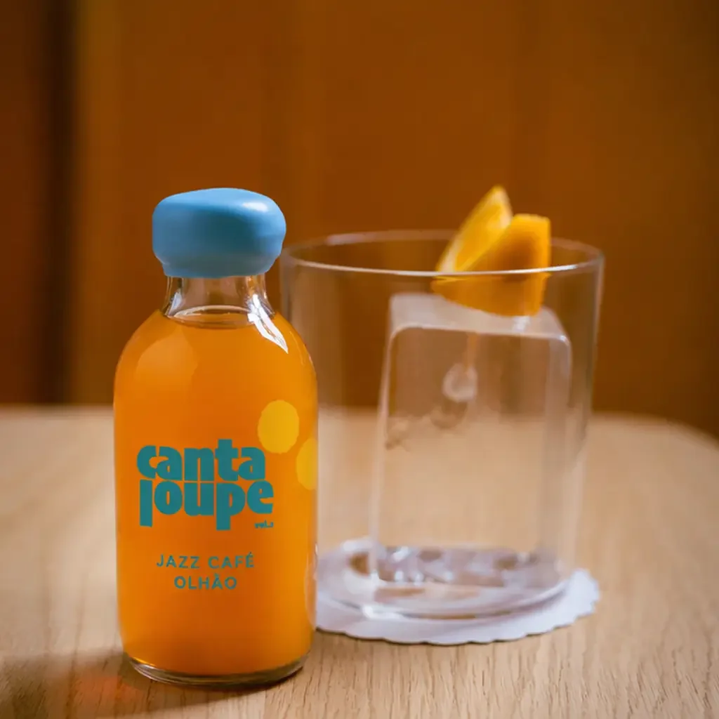 A small glass bottle with orange juice and a blue cap, labeled 'Canta Joue Jazz Café Olhão,' next to a glass filled with water, ice, and lemon slices on a wooden table.