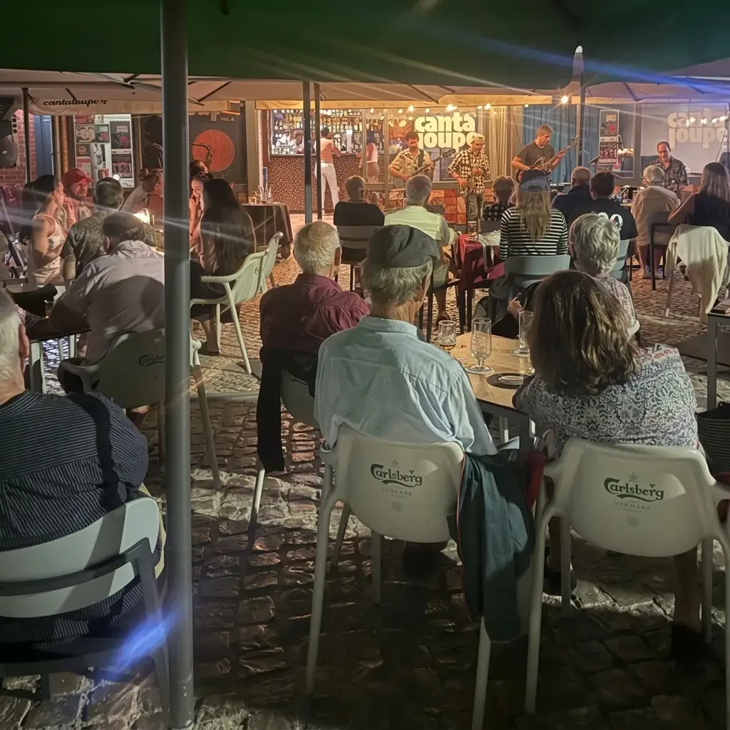 People watching a live outdoor music performance at a bar or restaurant during the evening. The stage has a band playing instruments under a tent with a sign that reads 'canta soupe'.