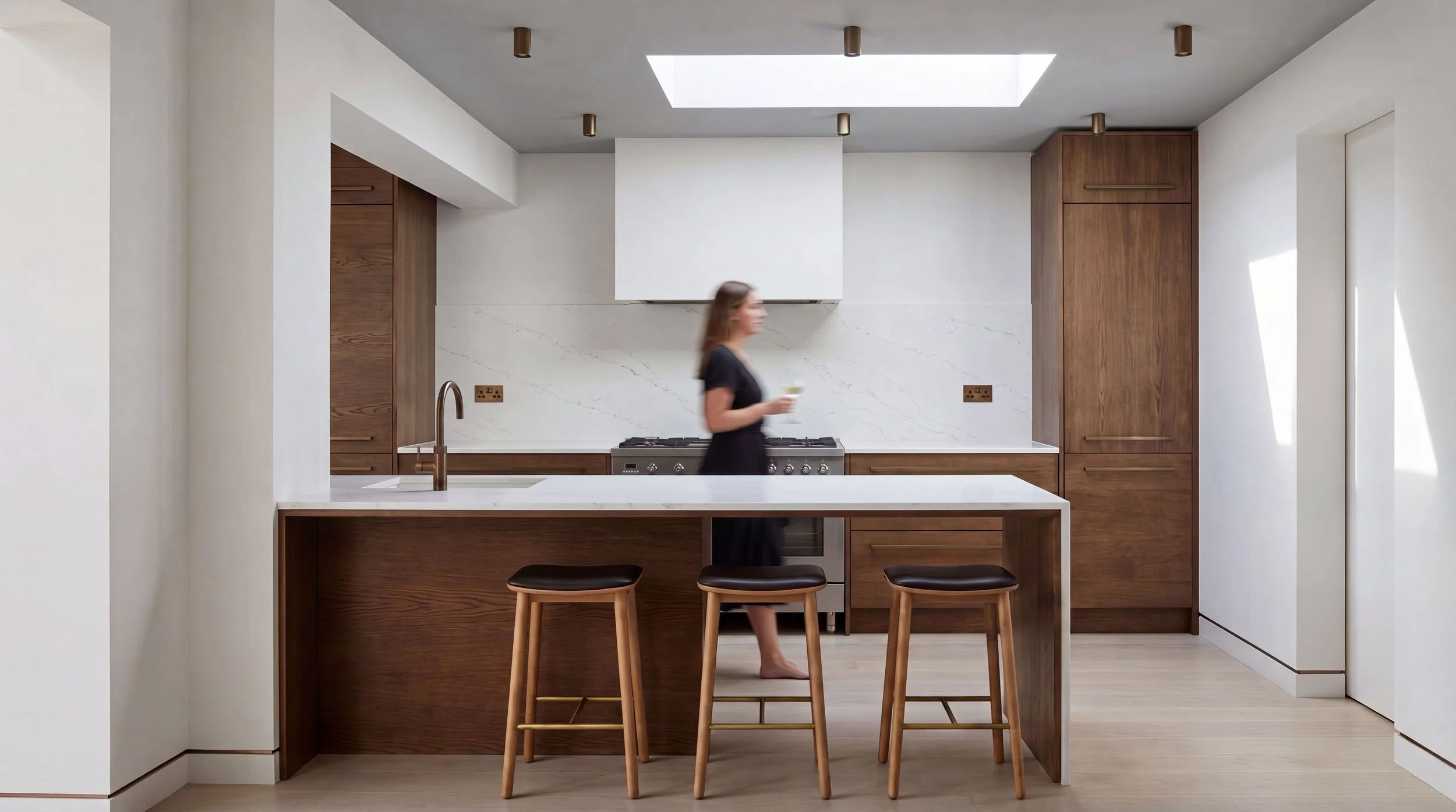 Modern kitchen with wooden cabinets, white marble backsplash, and an island with stools. A woman is walking with a drink.