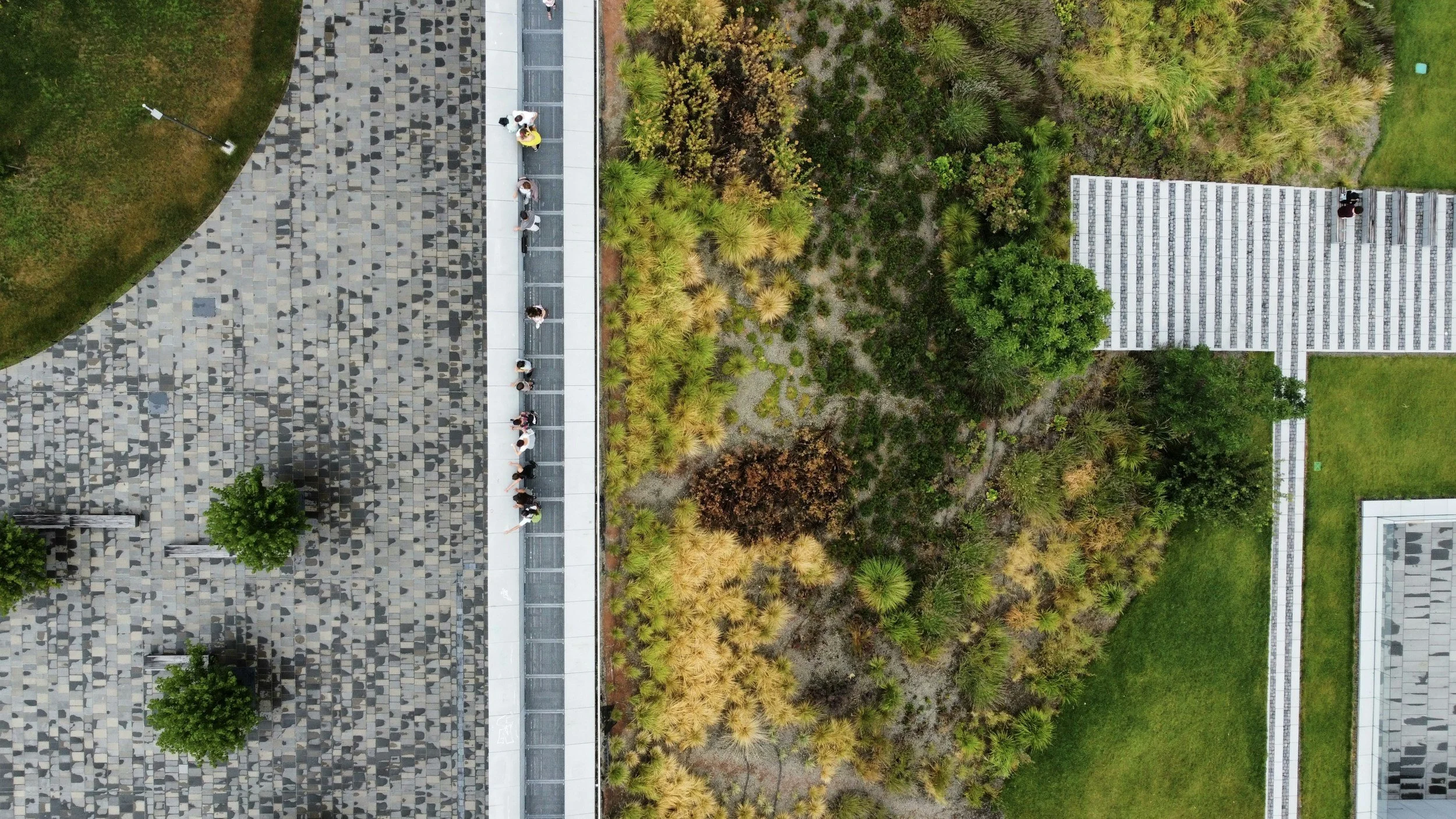 An aerial view of a modern building with a stone patterned roof, adjacent to a landscaped garden with various shrubs and trees. Several people are walking along a concrete pathway next to the building.