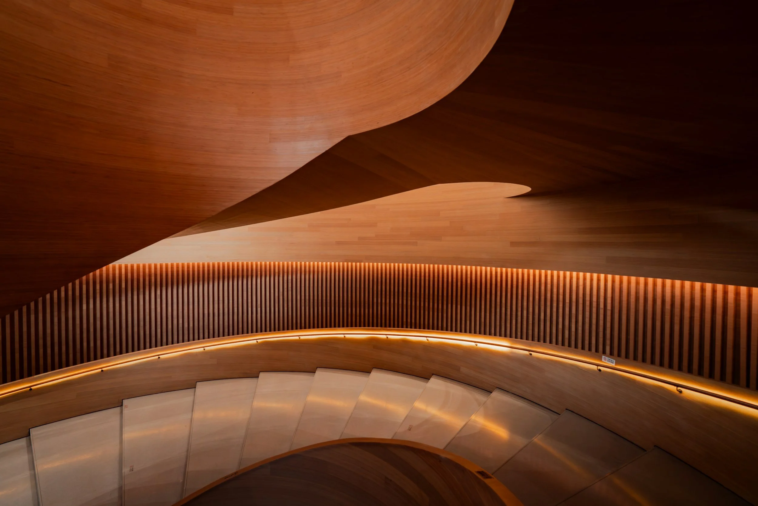 Curved wooden staircase with warm lighting on the handrail and vertical wooden slats on the wall.
