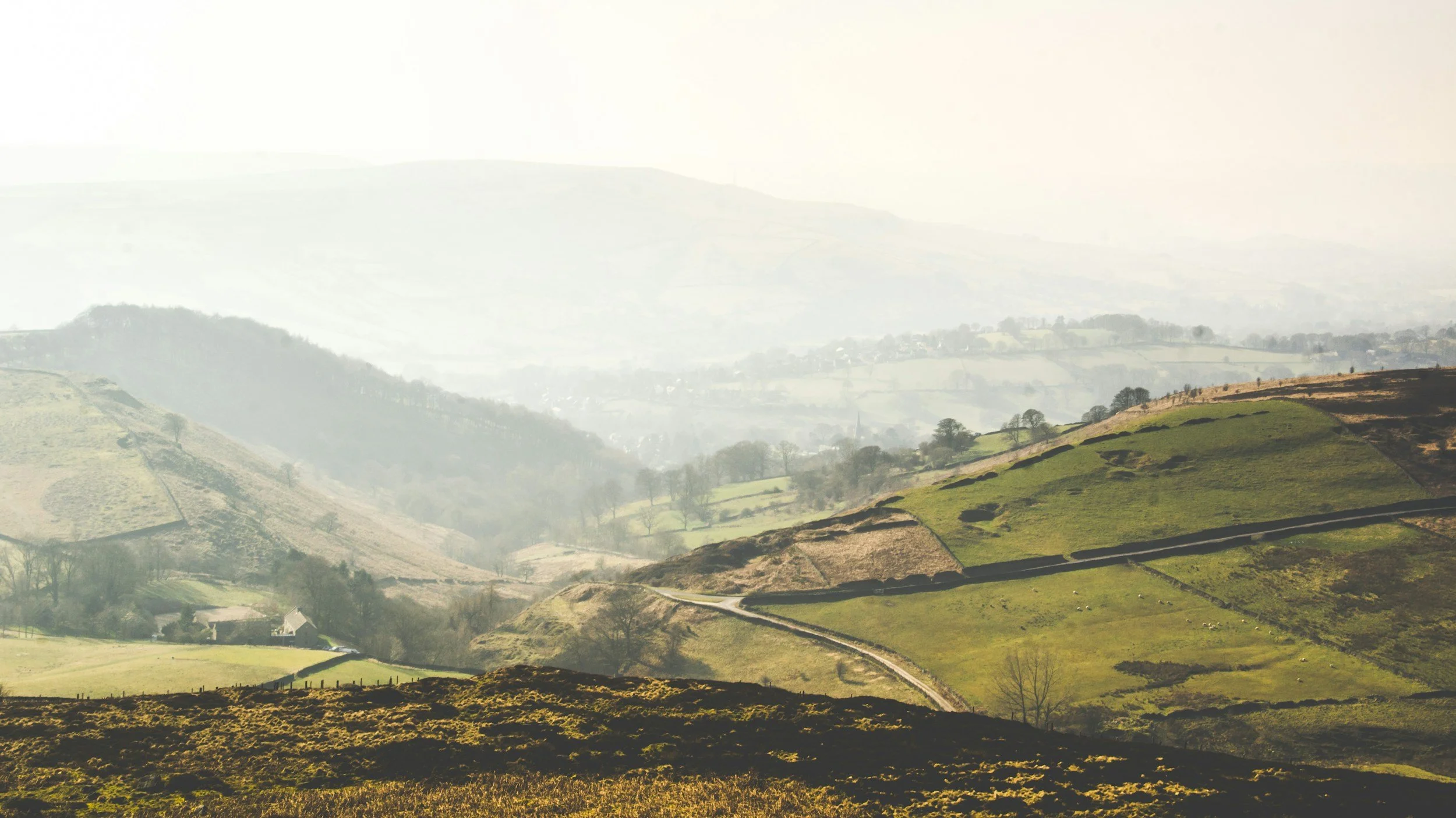 Scenic view of rolling green hills with patchwork agricultural fields and a few scattered houses, misty mountains in the background under a light cloudy sky.