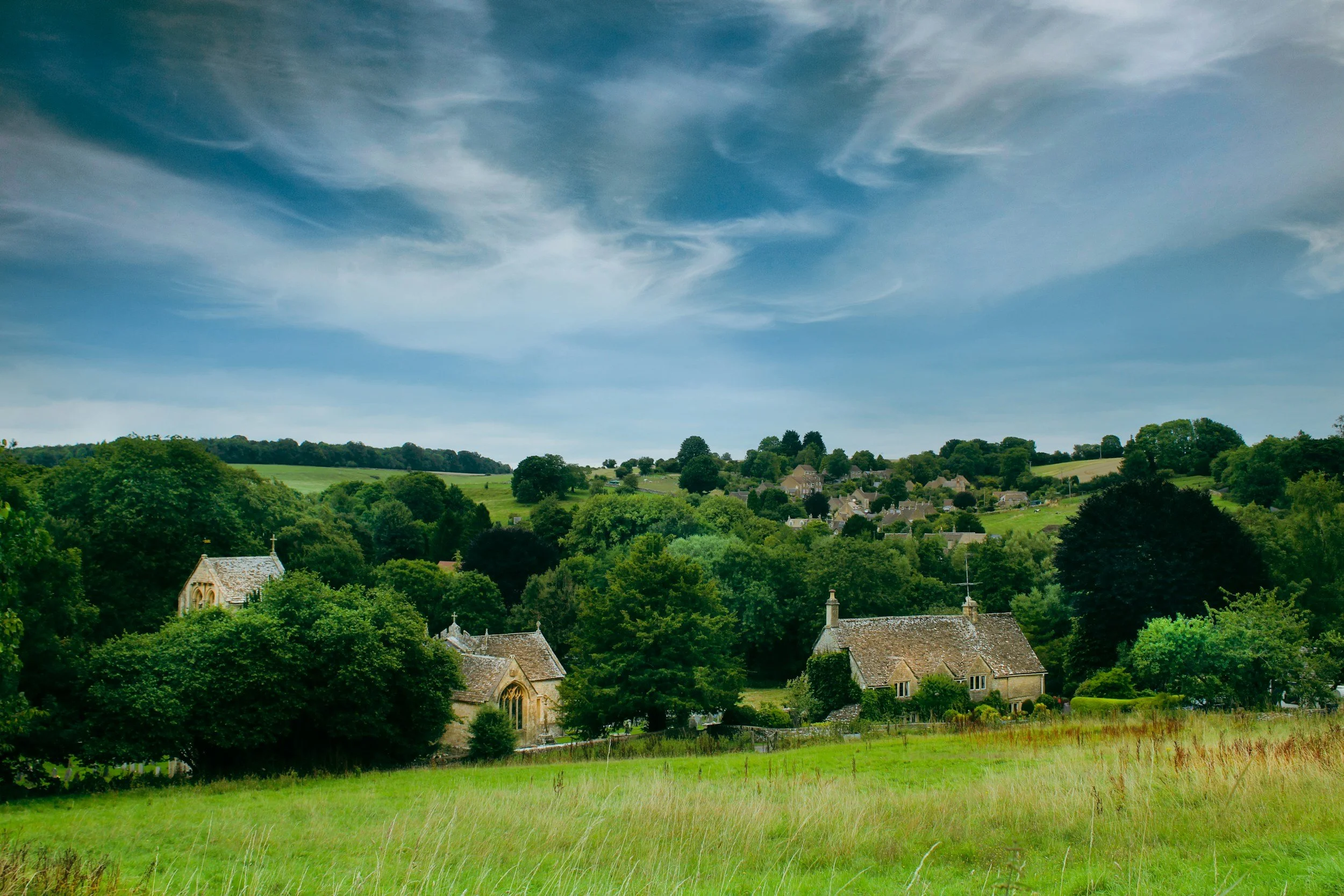 A scenic view of a small village in a lush green landscape with rolling hills, trees, and a church, under a partly cloudy sky.
