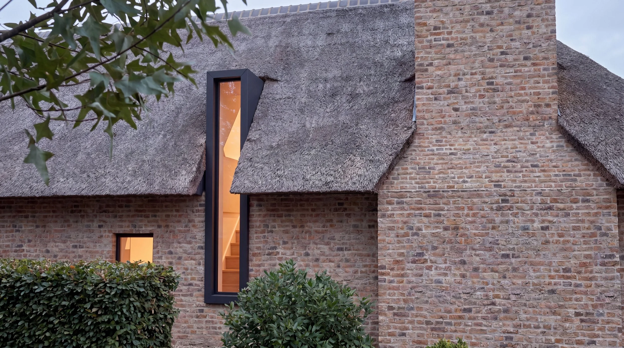 A modern house with brick walls, a thatched roof, a tall narrow window, and some green bushes in front.