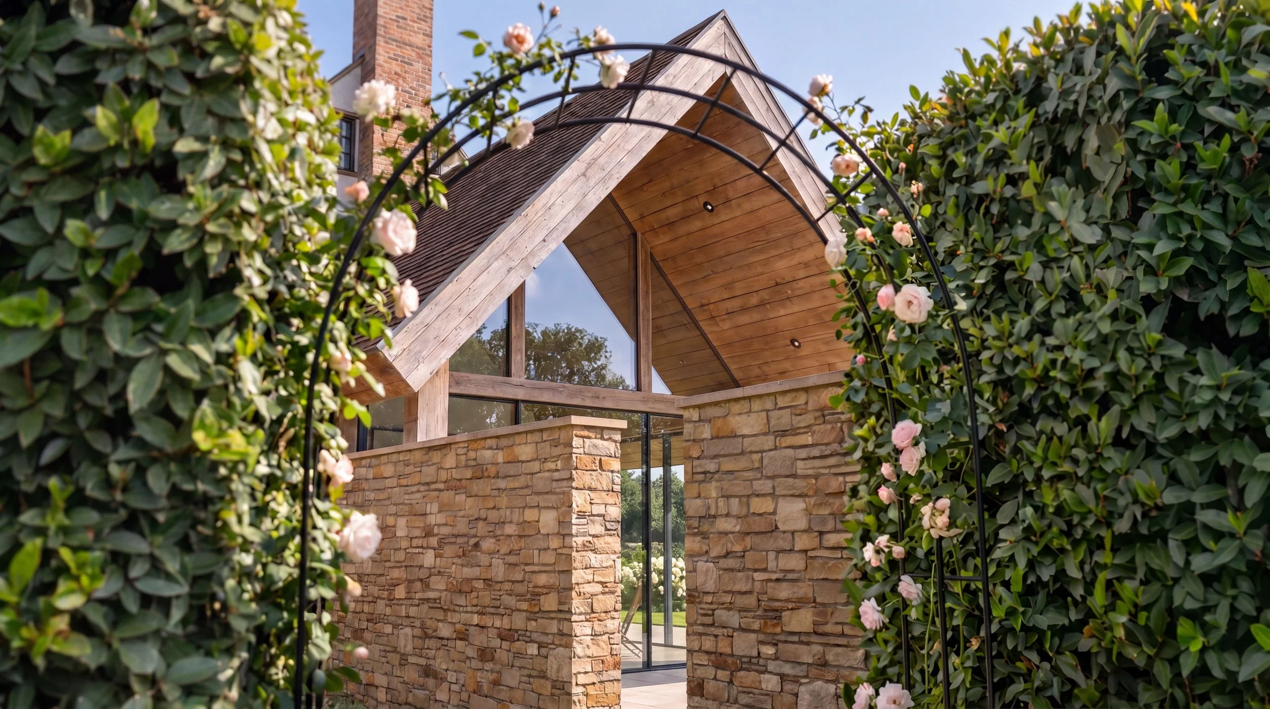 A modern house with a wooden roof, stone walls, and large glass windows surrounded by green bushes with pink flowers.