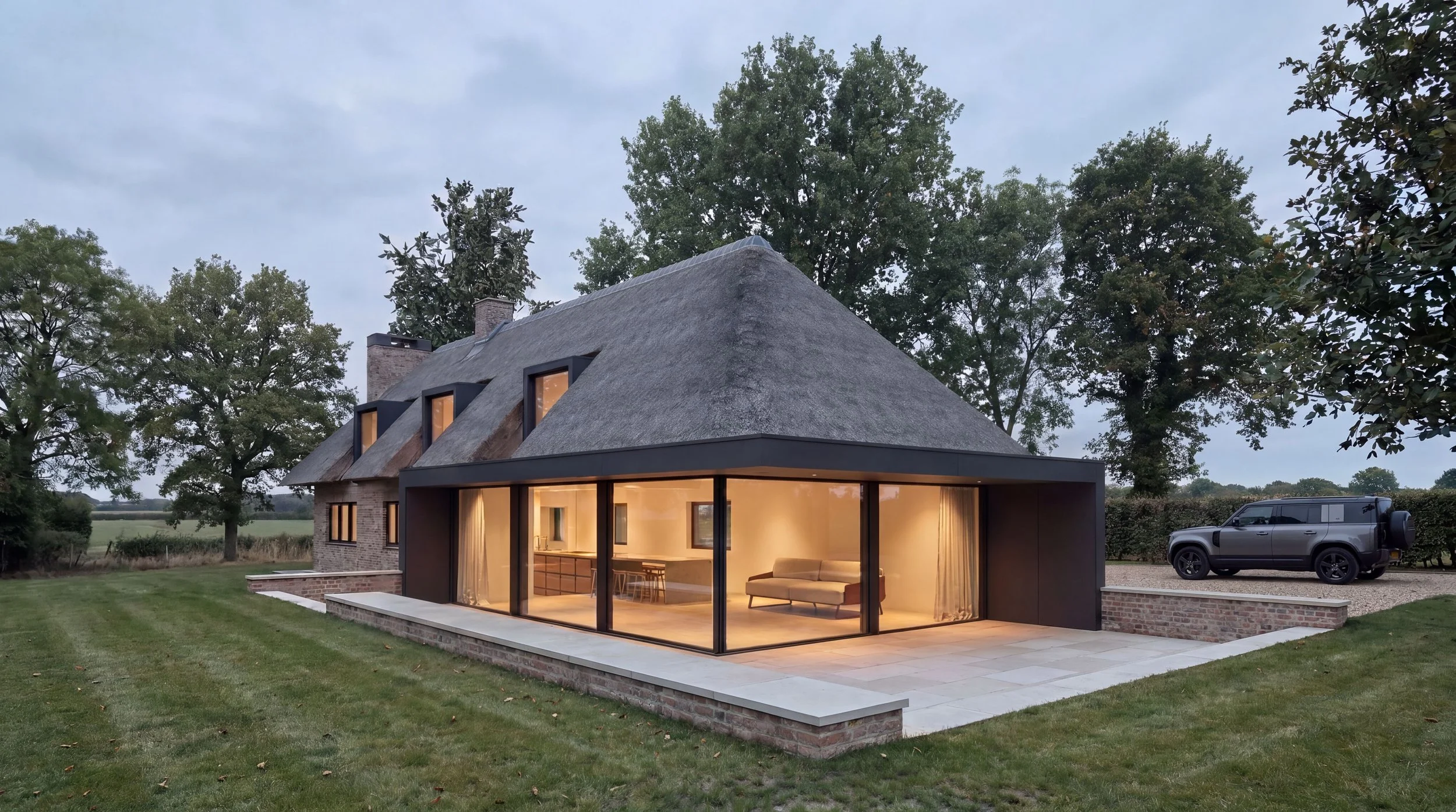 A modern house with a thatched roof, large glass windows, and a patio, surrounded by trees and a lawn, with a black vehicle parked nearby.
