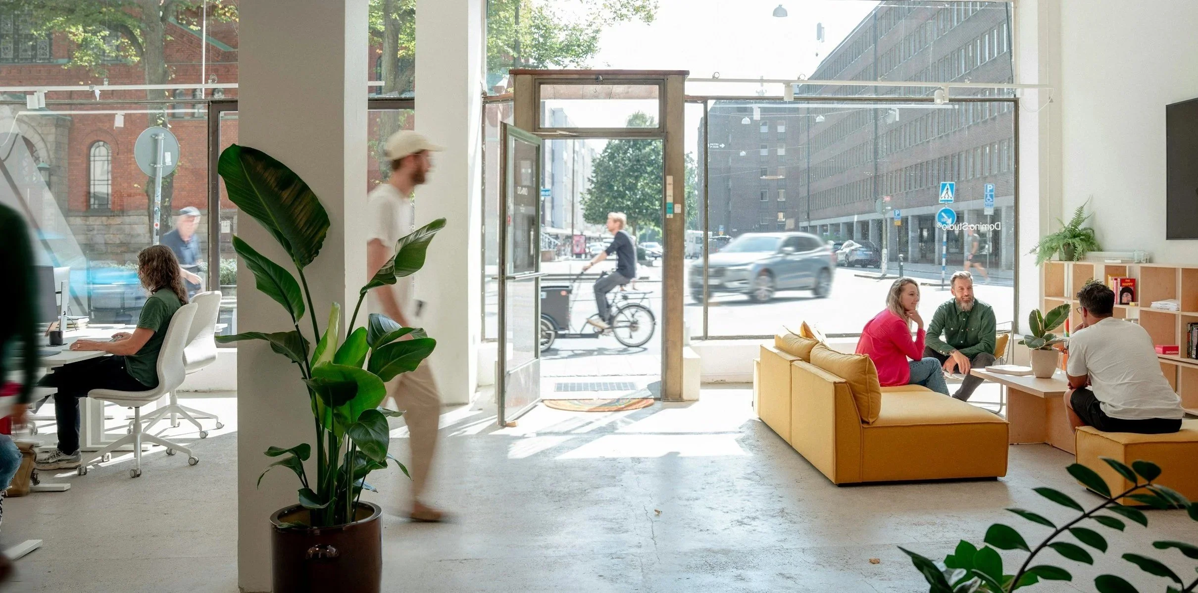 Interior of a modern coffee shop with large front windows showing street view, several customers sitting and talking, a person working at a desk, and a man cycling outside.