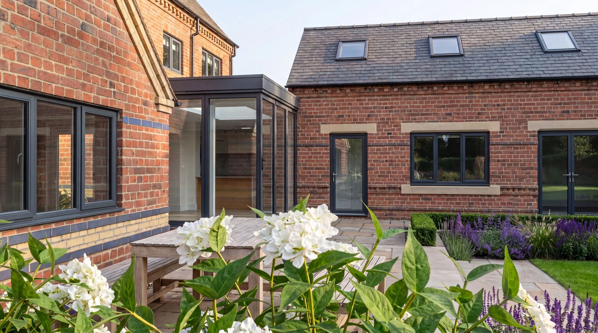 A backyard scene with a brick house featuring a glass sunroom, large windows, and a landscaped garden with flowering plants, bushes, and a wooden deck.