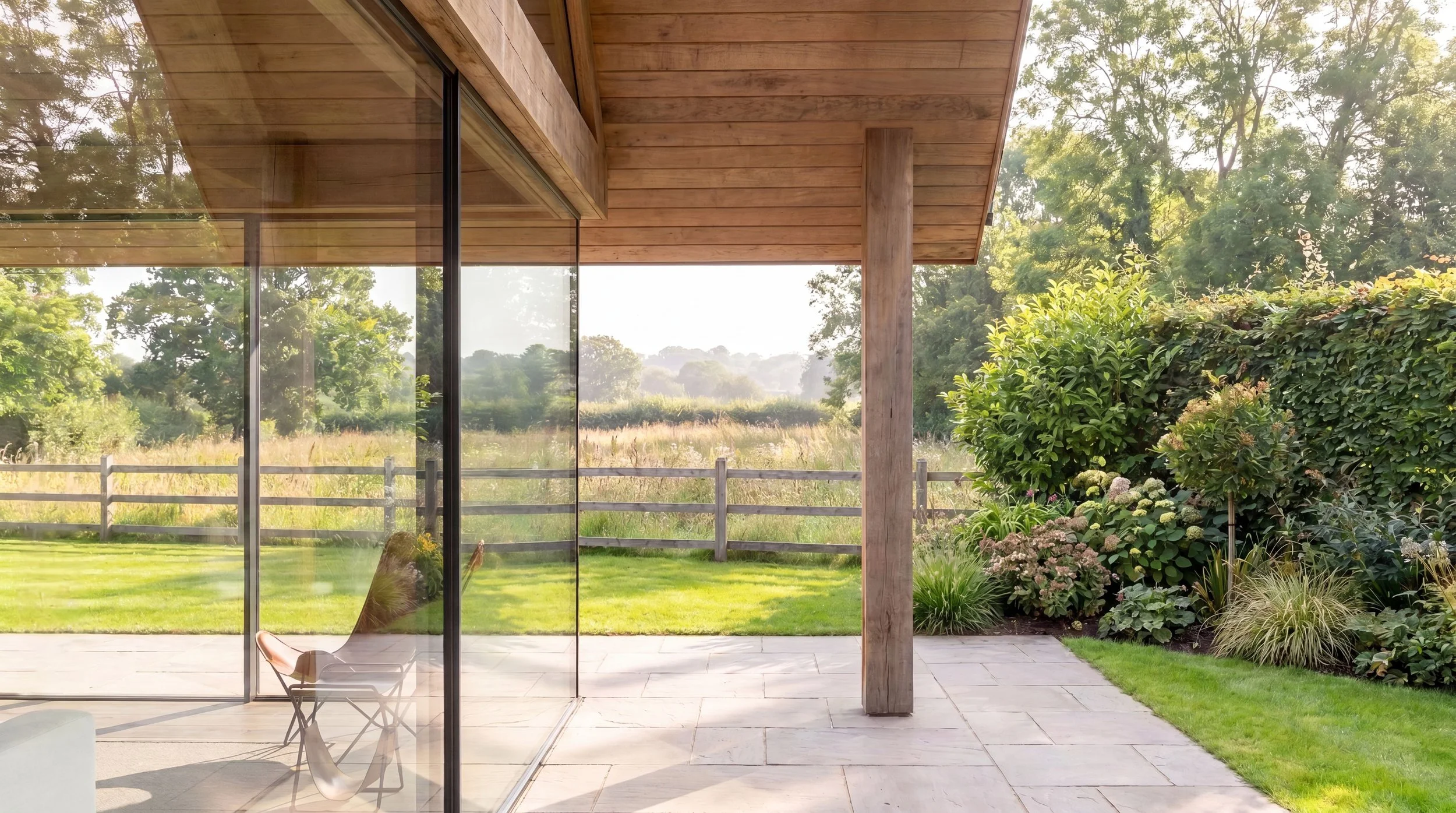View from inside a modern house with large glass sliding doors, showing a backyard with a grassy lawn, a wooden fence, trees, bushes, and a patio area.