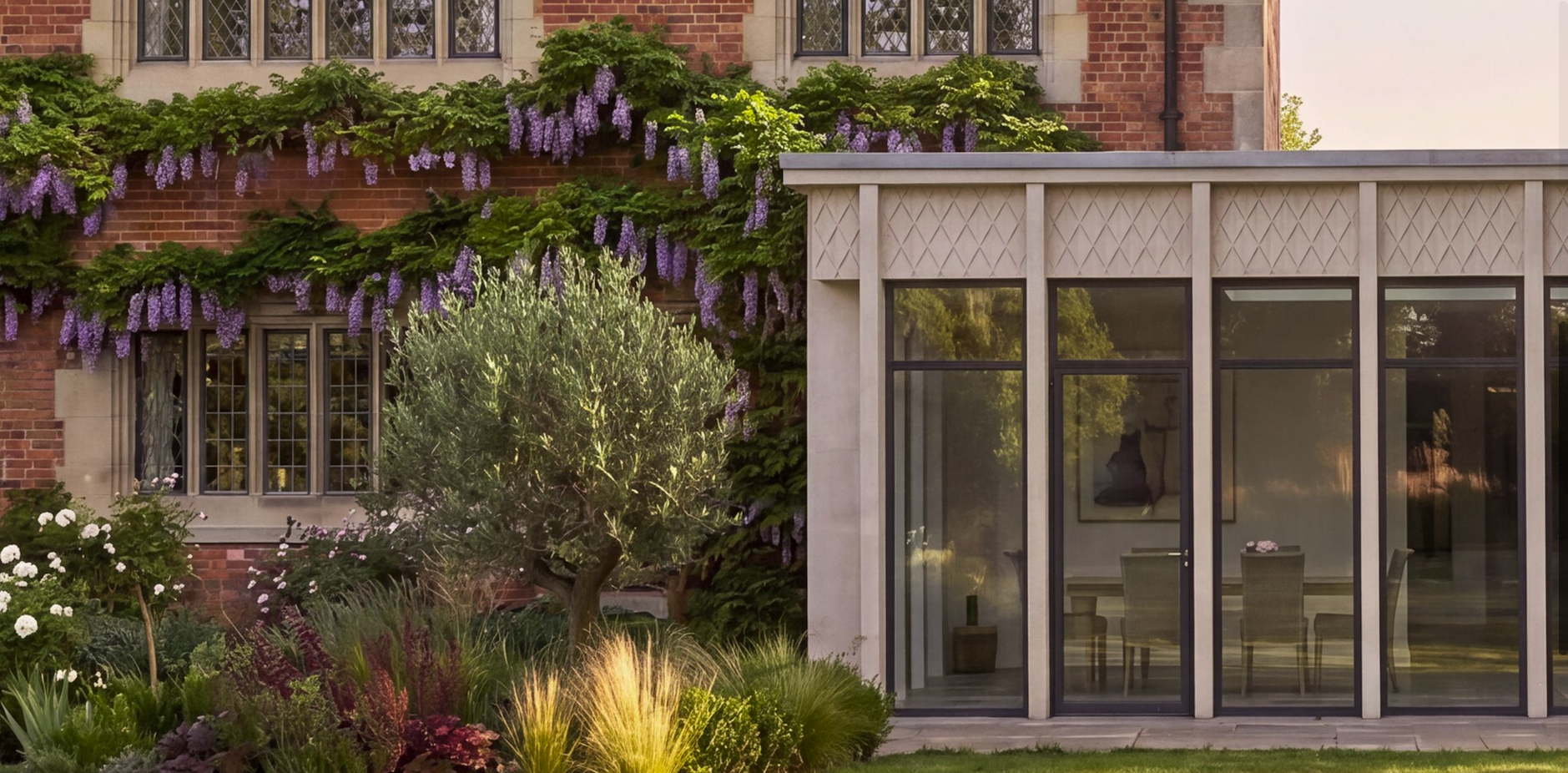 A garden with lush greenery, flowering vines on a brick house, and a modern glass-walled extension with a dining area inside.