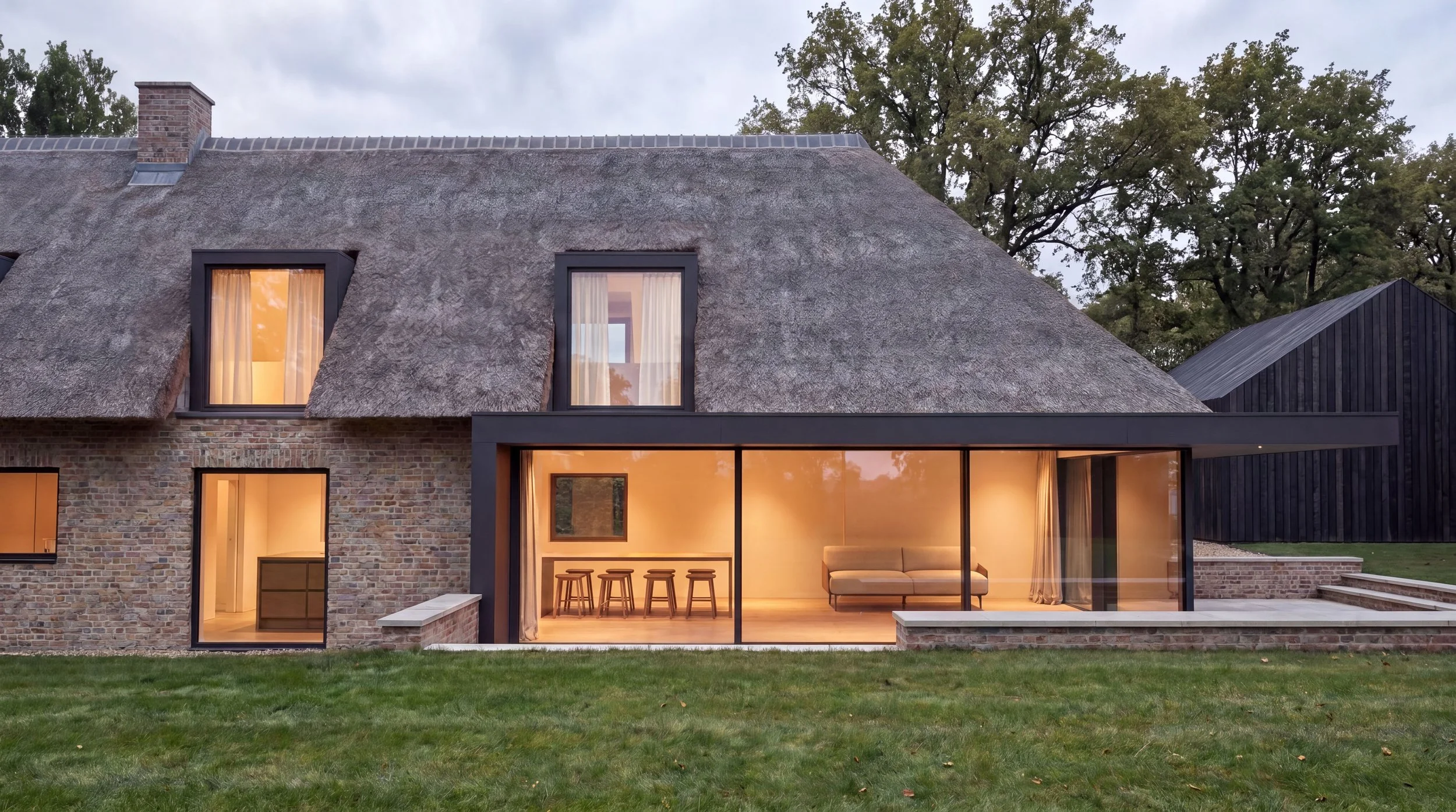 Modern house with a thatched roof, brick lower section, large glass doors, and windows illuminated from inside, surrounded by a lawn and trees in the background.