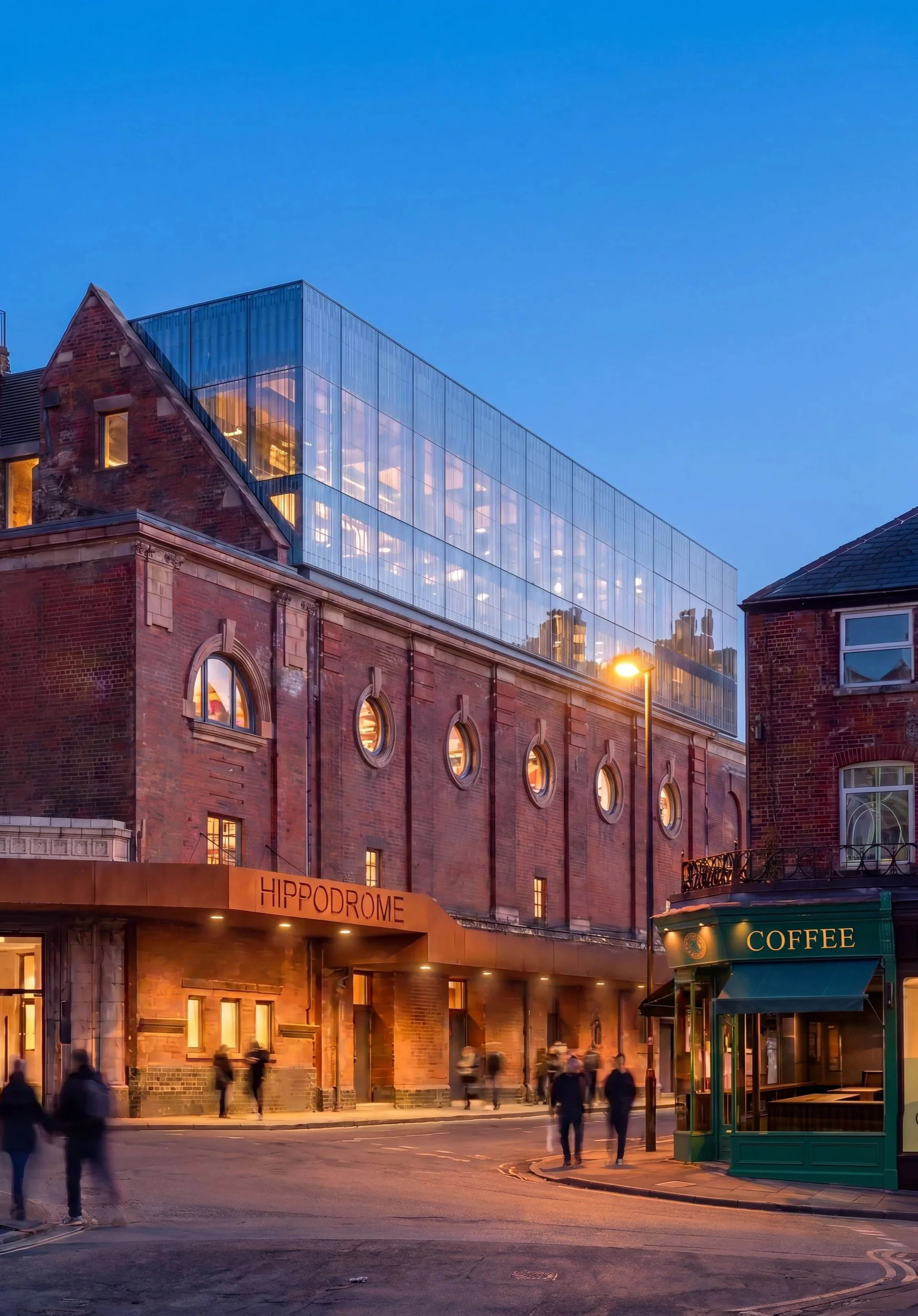 A city street scene at dusk with a red brick building featuring a modern glass extension on top, illuminated windows, street lamps, and a small green coffee kiosk, with people walking on the sidewalk.