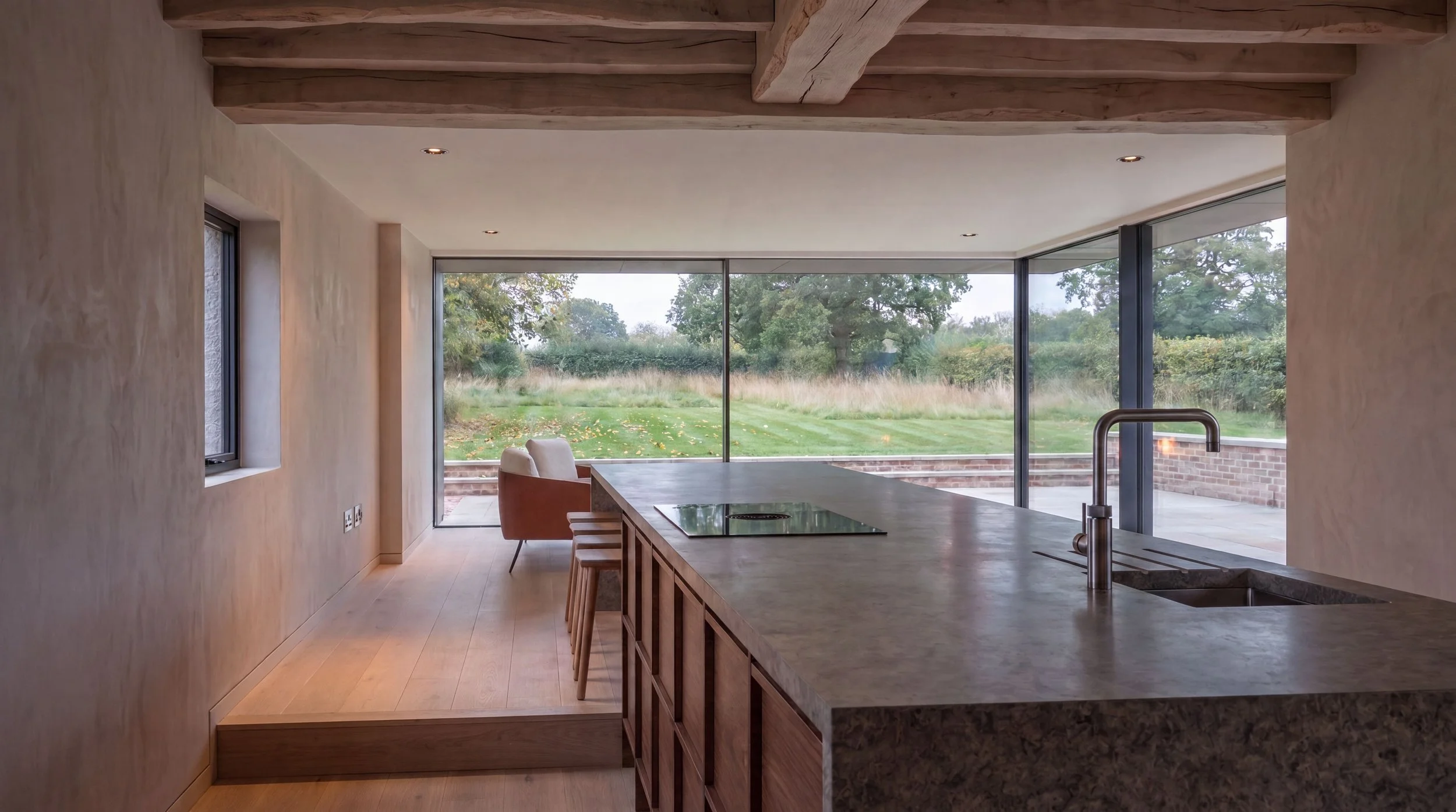 A modern kitchen with a large island, natural stone worktop, wooden stools, and a view of a green outdoor landscape through floor-to-ceiling glass doors.