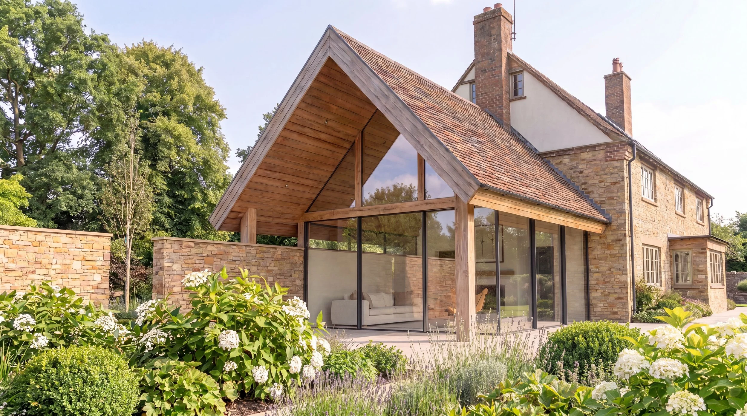 A modern house with a stone exterior and a sloped roof, featuring large glass windows and a wooden patio area, surrounded by lush green trees and flowering shrubs.