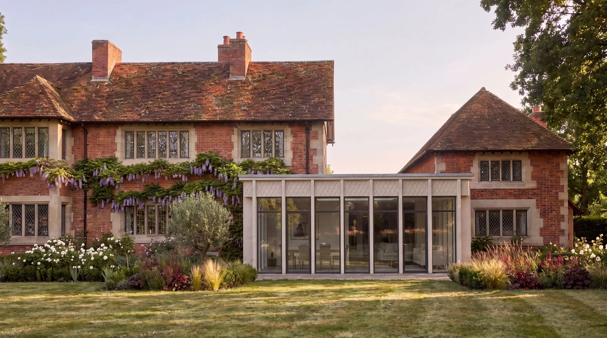 A large brick house with a sloped tiled roof, multiple chimneys, and large windows. A modern glass extension is attached on the right side of the house. There are lush gardens with flowering plants and trees in front of the house and a green lawn.