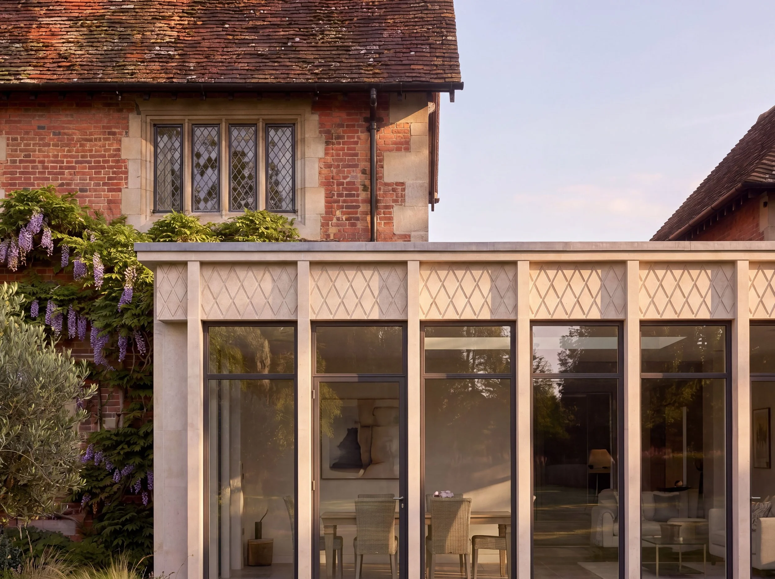 A red brick manor house with a beautiful stone and glass conservatory extension. There are purple flowering plants and greenery in front.