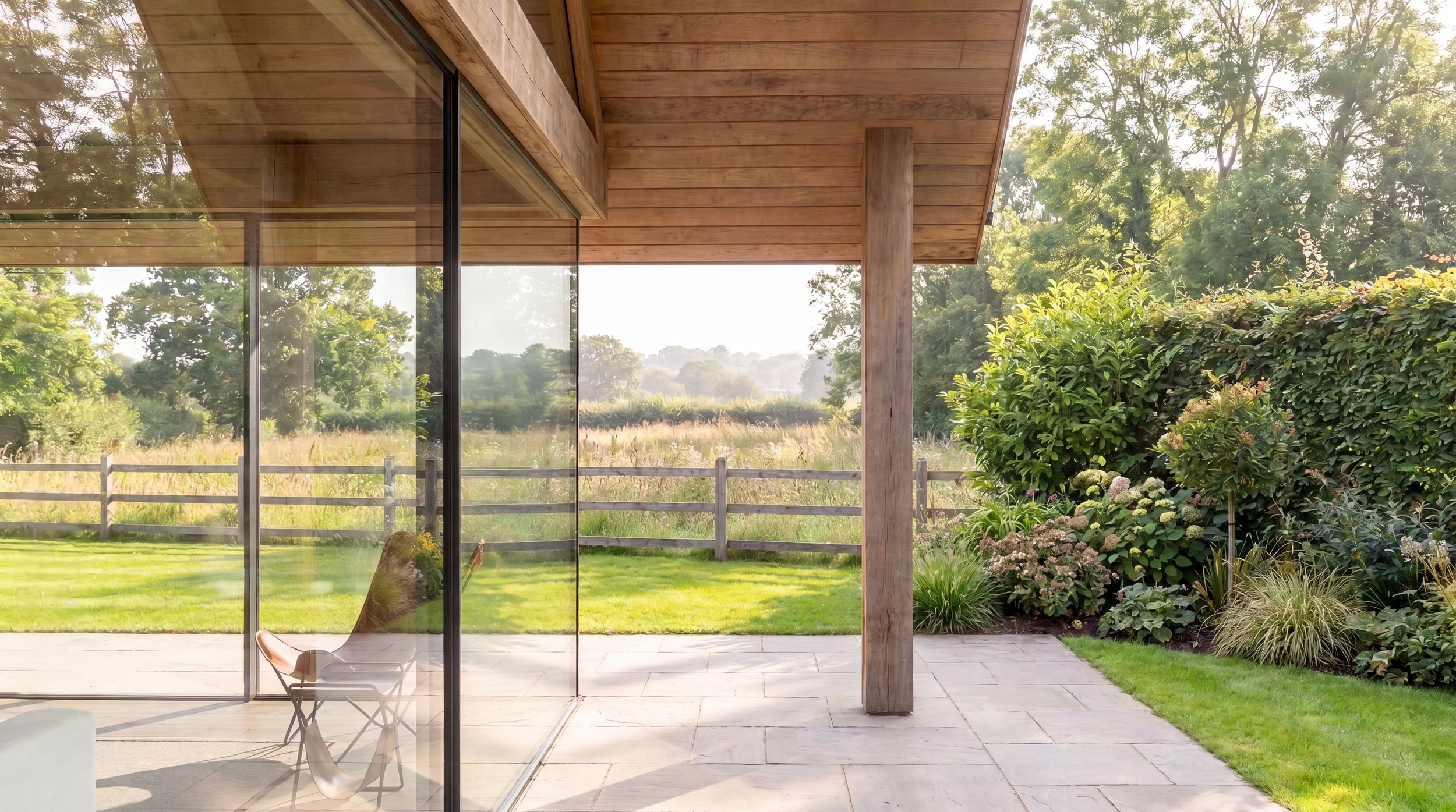 A beautifully minimalist modern house extension with large glass sliding doors, oak column and cladding, and natural landscape including trees, bushes and meadows.