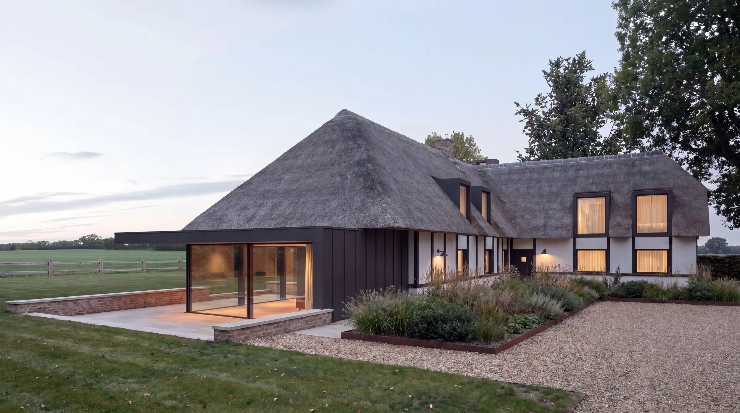 Modern house with a thatched roof, large windows with curtains, and a garden with plants and gravel path, set in a rural landscape at dusk.
