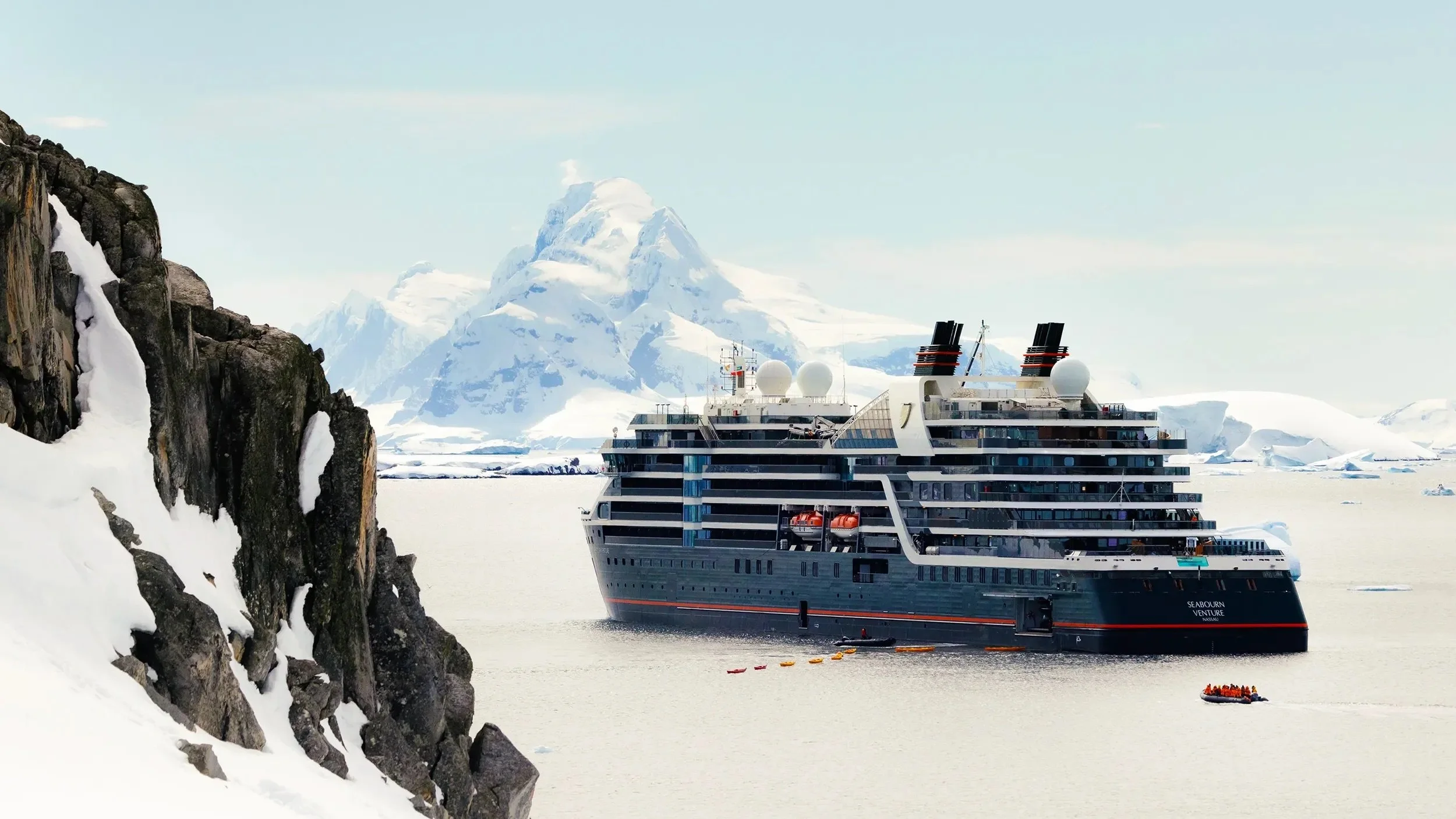 A large cruise ship named Seabourn Venture with two exhaust stacks and satellite domes, floating in icy waters near snow-covered rocky cliffs and a snow-capped mountain in the background.