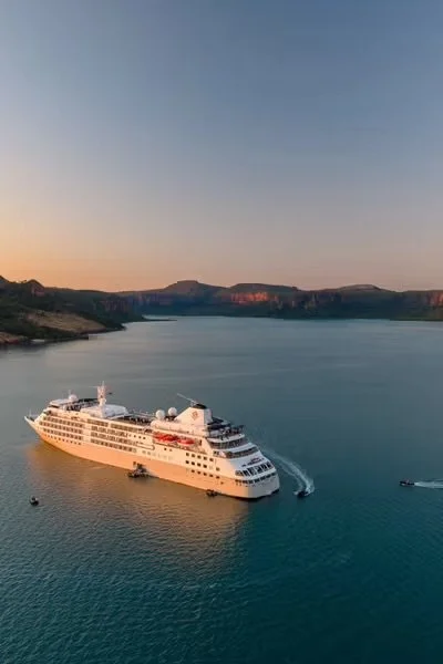 A large white cruise ship sailing on a calm body of water during sunset, with hills in the background.