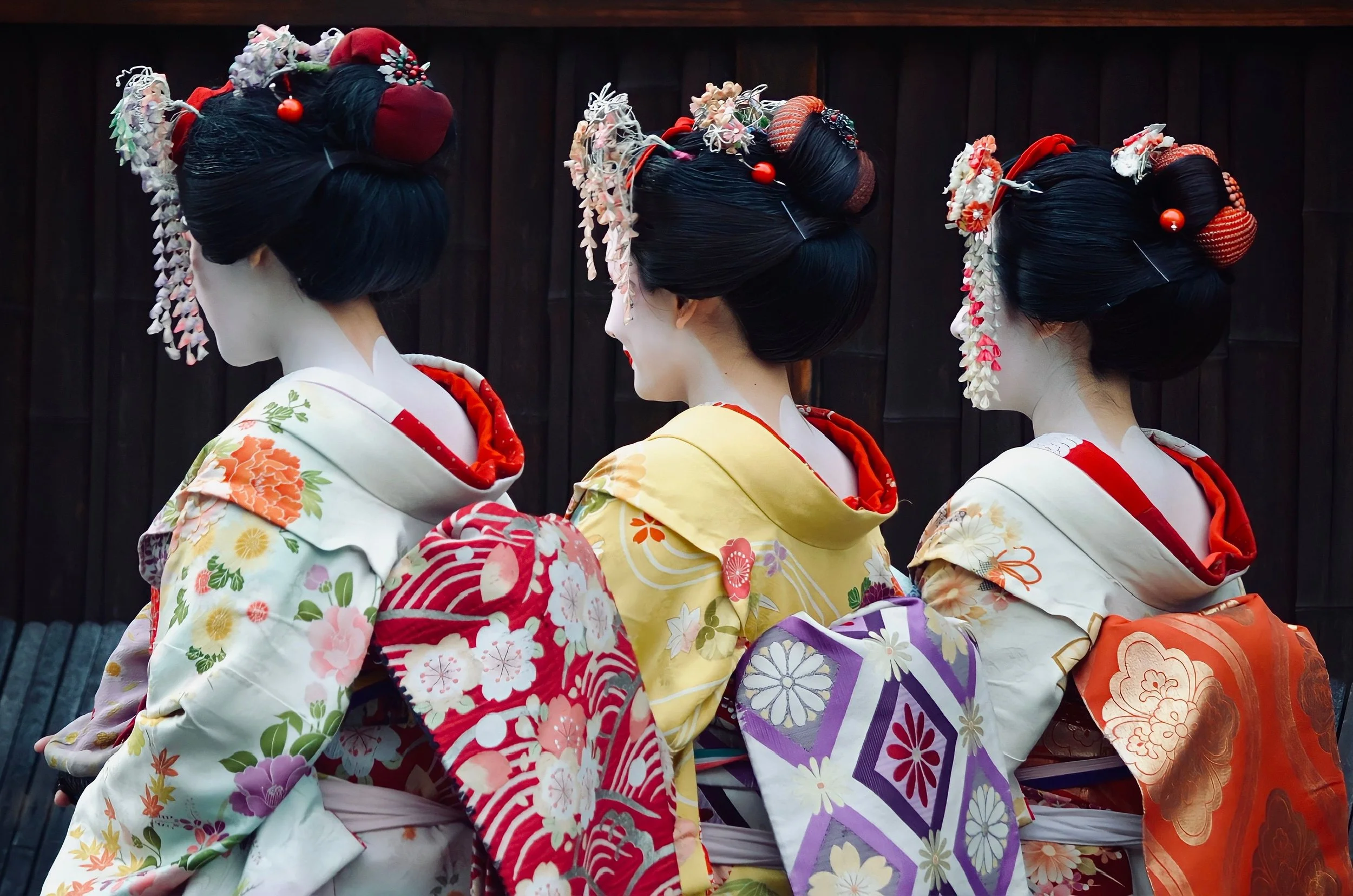 Three women dressed as geishas in colorful kimonos with elaborate hairstyles and hair accessories, standing side by side against a dark background.