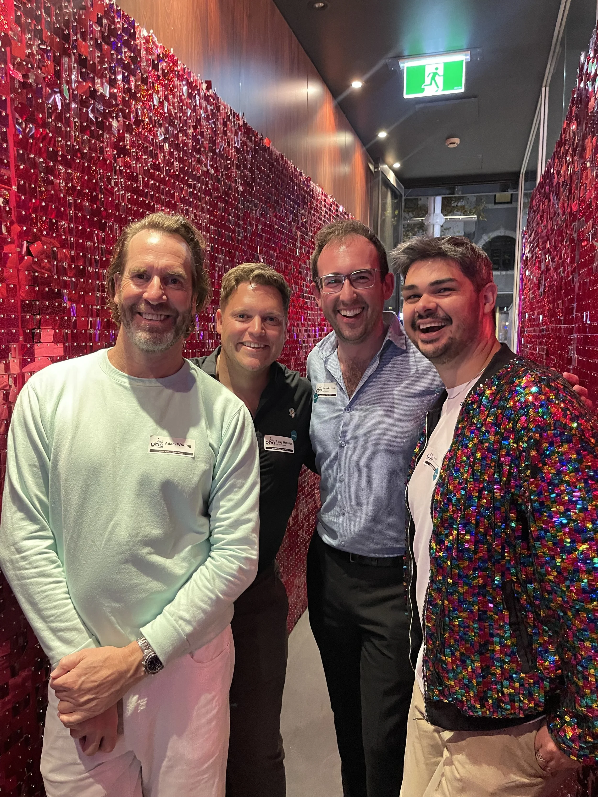 Four men standing together in front of a red decorative wall, smiling at the camera. They are wearing casual or semi-formal clothing, with name tags visible on two of them. The setting appears to be indoors, possibly at a social or professional event.