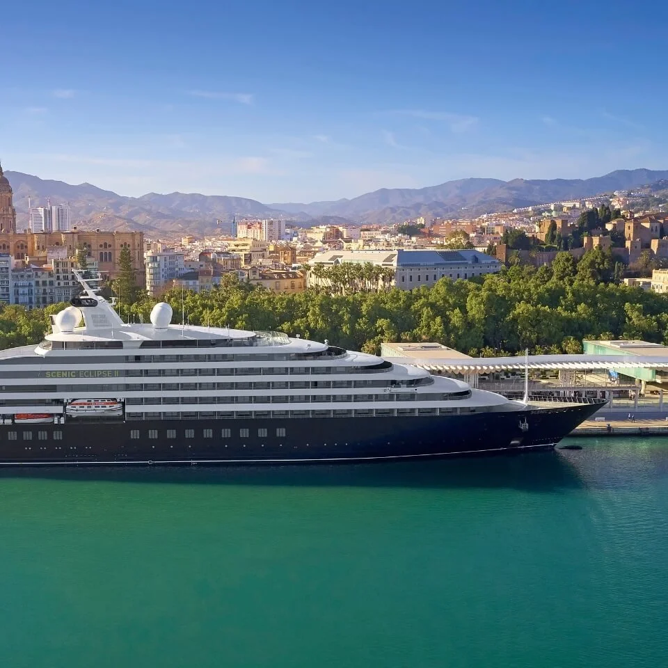 Large luxury yacht docked in a harbor with a city and mountainous landscape in the background under a clear blue sky.