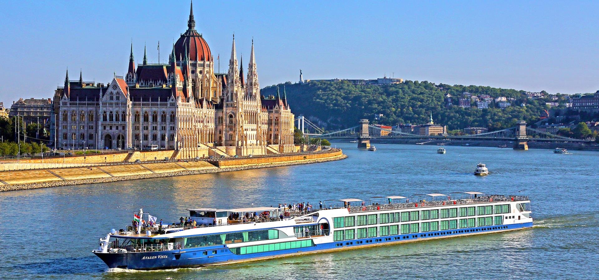 A large river cruise boat named Avalon Vista sailing on the Danube River near the Hungarian Parliament Building in Budapest, with a cityscape and hills in the background.