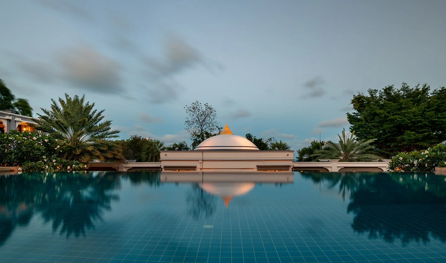 Reflection of a white-domed building with a golden top and surrounding greenery in a pool at sunset.