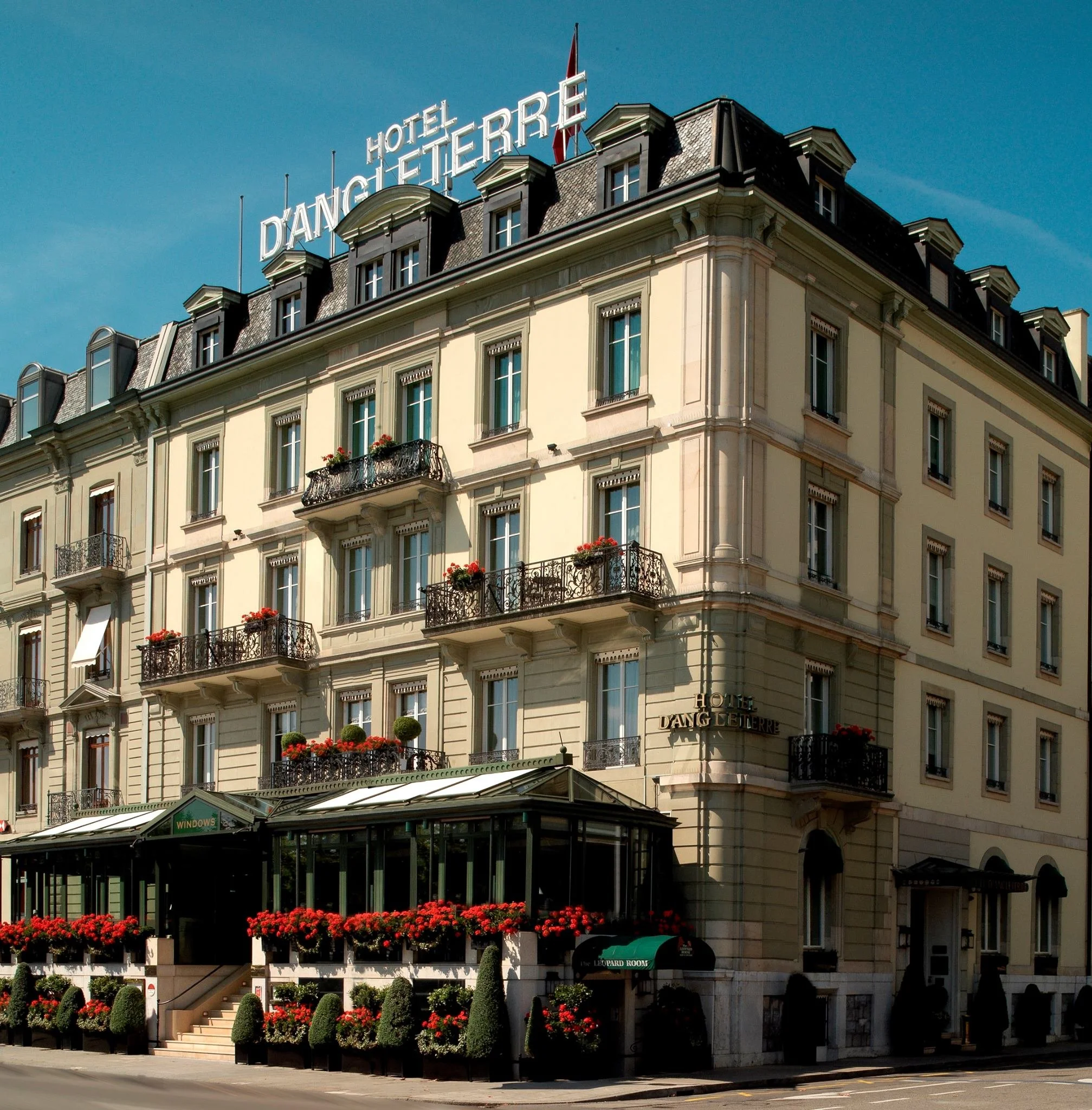 Fifth floor of a hotel building with a sign reading 'Hotel D'ANGHEITERRRE' and decorative windows, balconies with flower boxes, and a glass conservatory at street level with red flowers in planters.