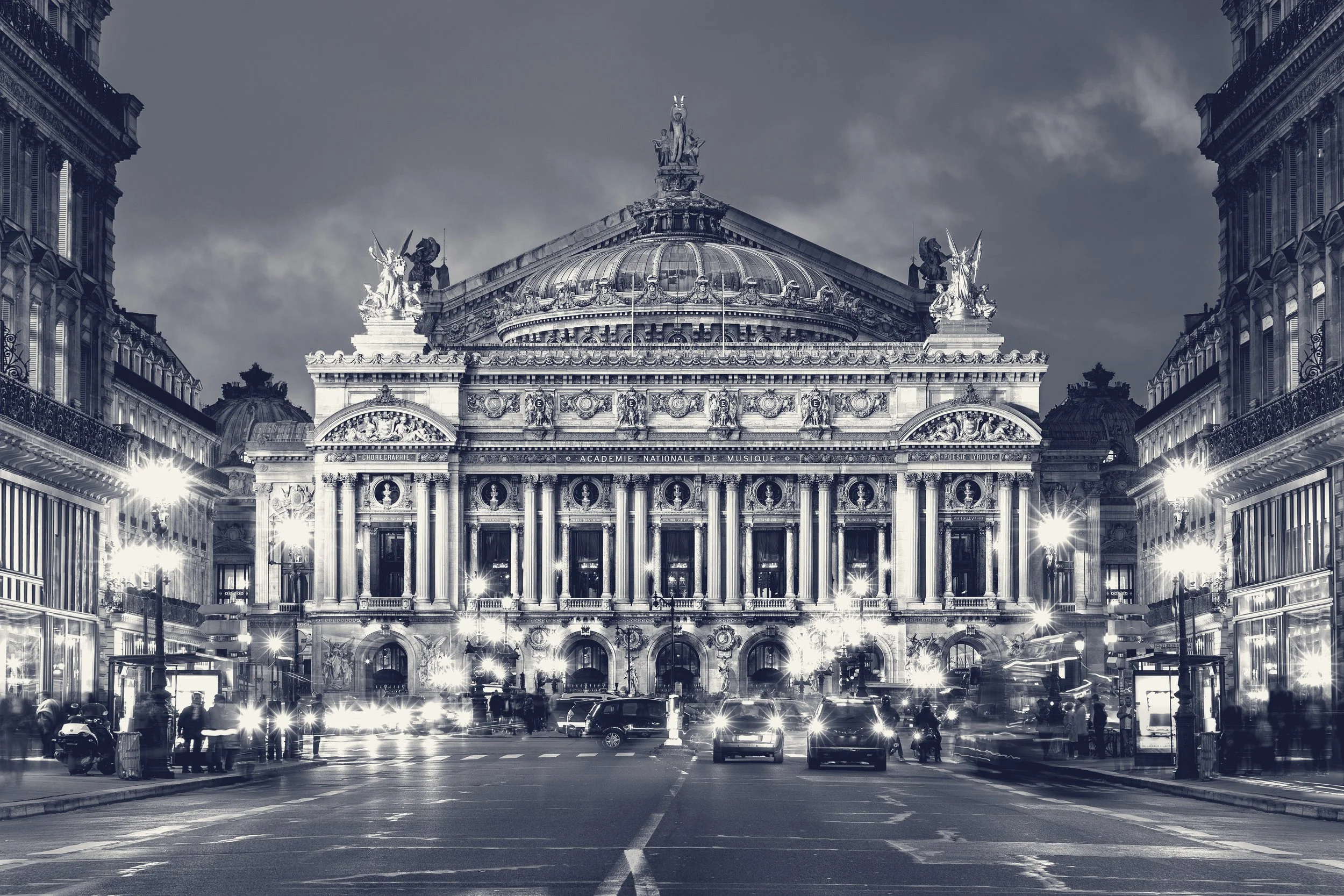 A large, ornate historic building with a domed roof and statues on top, illuminated at night, situated between narrower buildings with intricate facades. There are cars and pedestrians on the street in front of the building.
