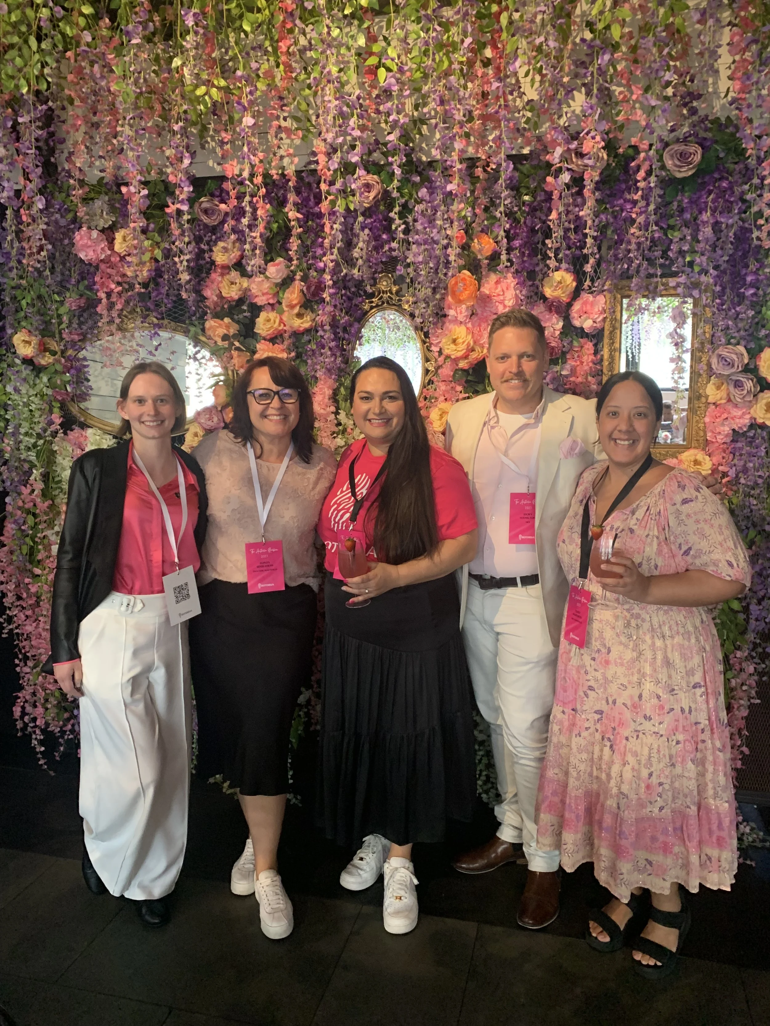 Group of five people standing in front of a floral wall with pink, purple, and yellow flowers and hanging vines. They are smiling, wearing conference badges around their necks, and holding drinks.