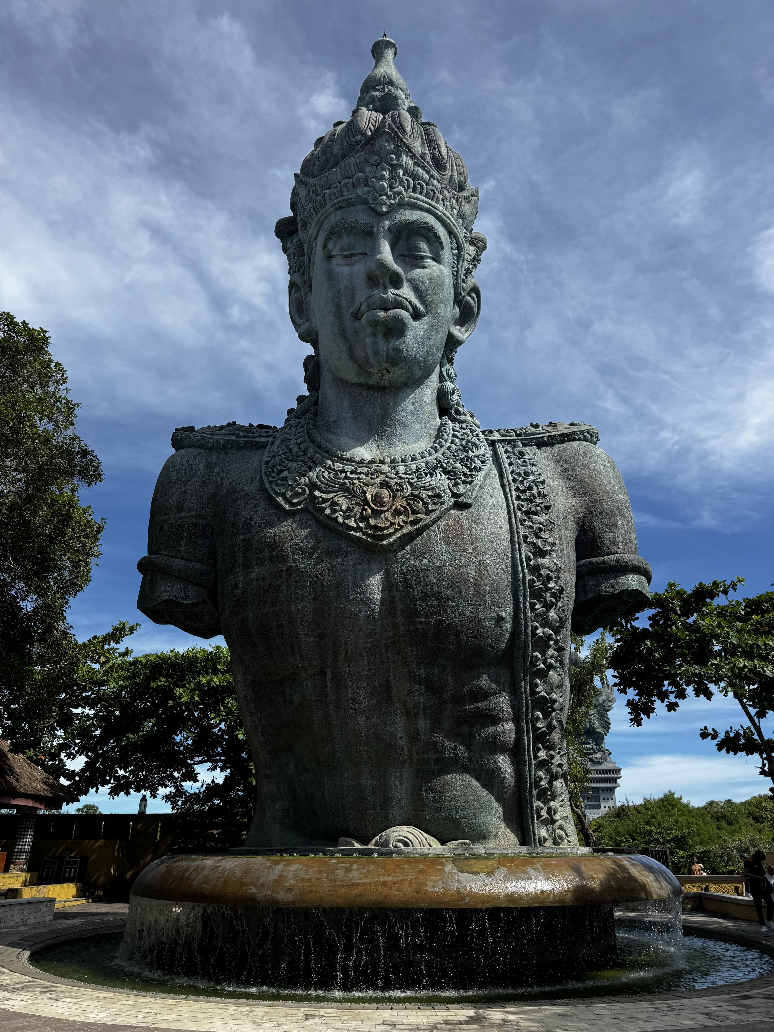 Statue of a deity with an ornate headdress, standing in a fountain surrounded by greenery.