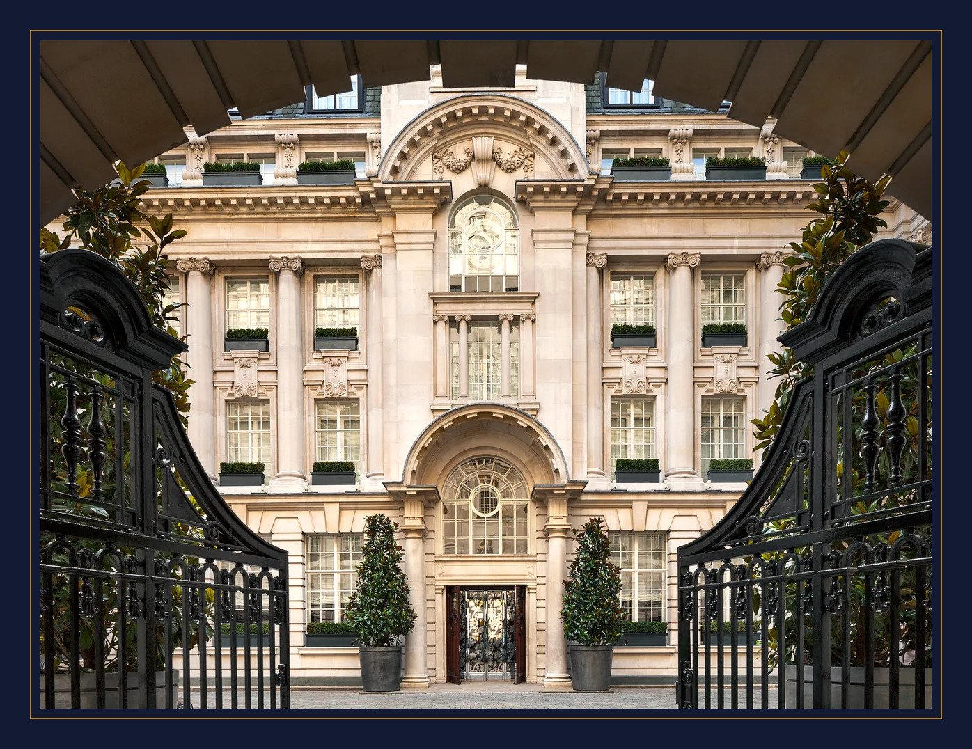Luxury building with classical architecture, large windows with shutters, columns, and decorative stonework, viewed through an open gate with plants and greenery.