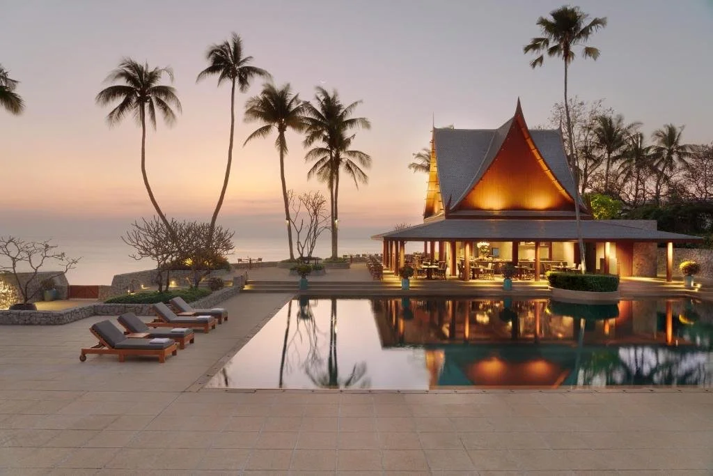 A luxurious seaside resort with a pool, palm trees, and a traditional Thai-style pavilion illuminated at sunset.