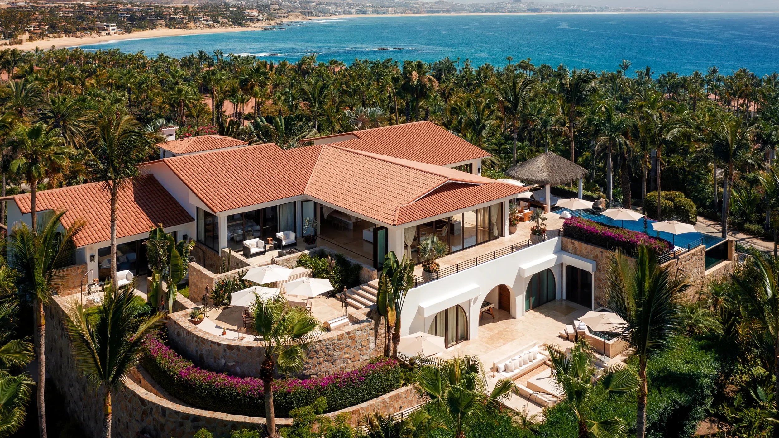 Aerial view of a luxurious beachfront property with white walls, terracotta roofs, a swimming pool, surrounded by lush greenery and palm trees, overlooking the ocean.
