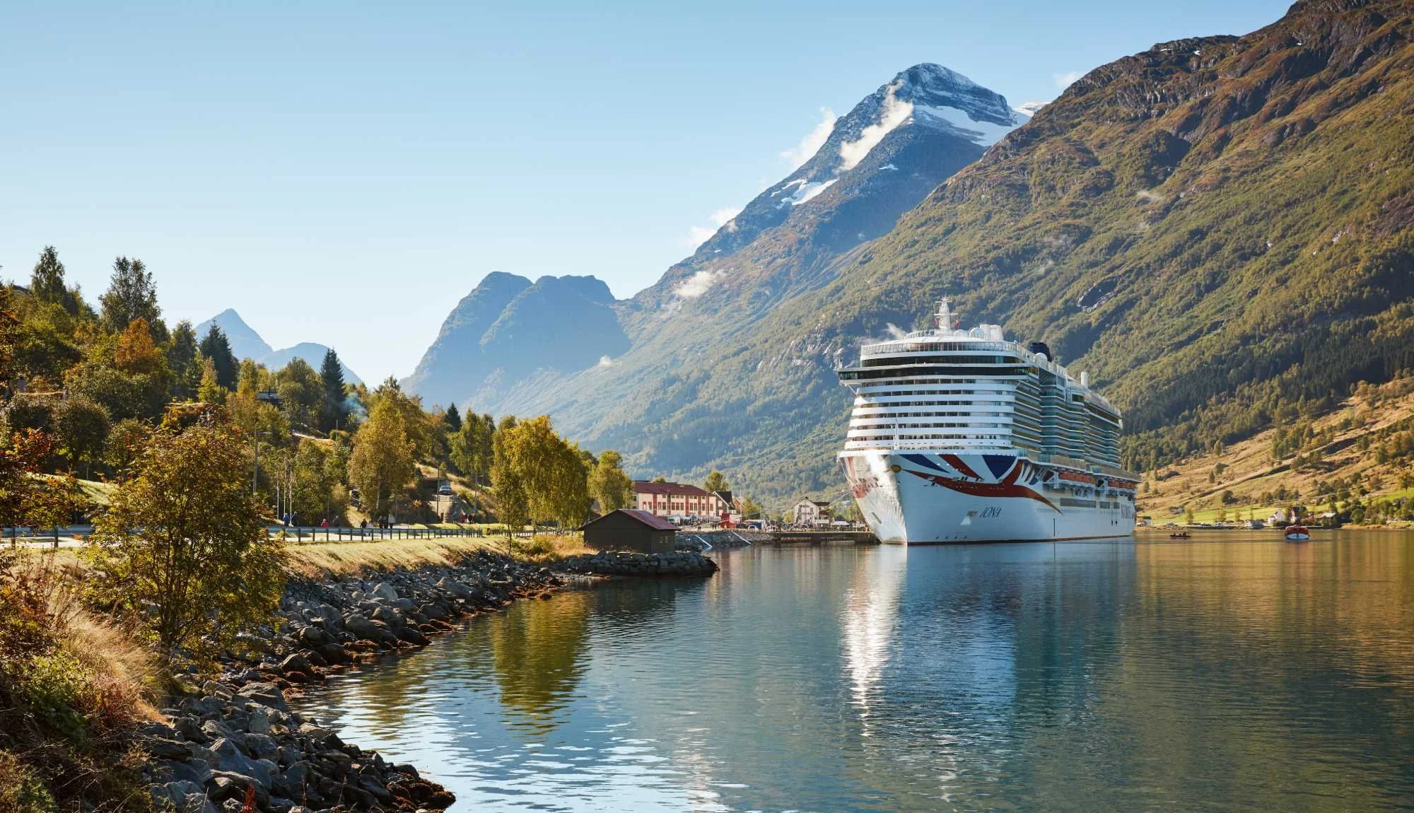 Large cruise ship docked in a narrow waterway surrounded by lush green trees, with tall mountain peaks in the background under a clear blue sky.