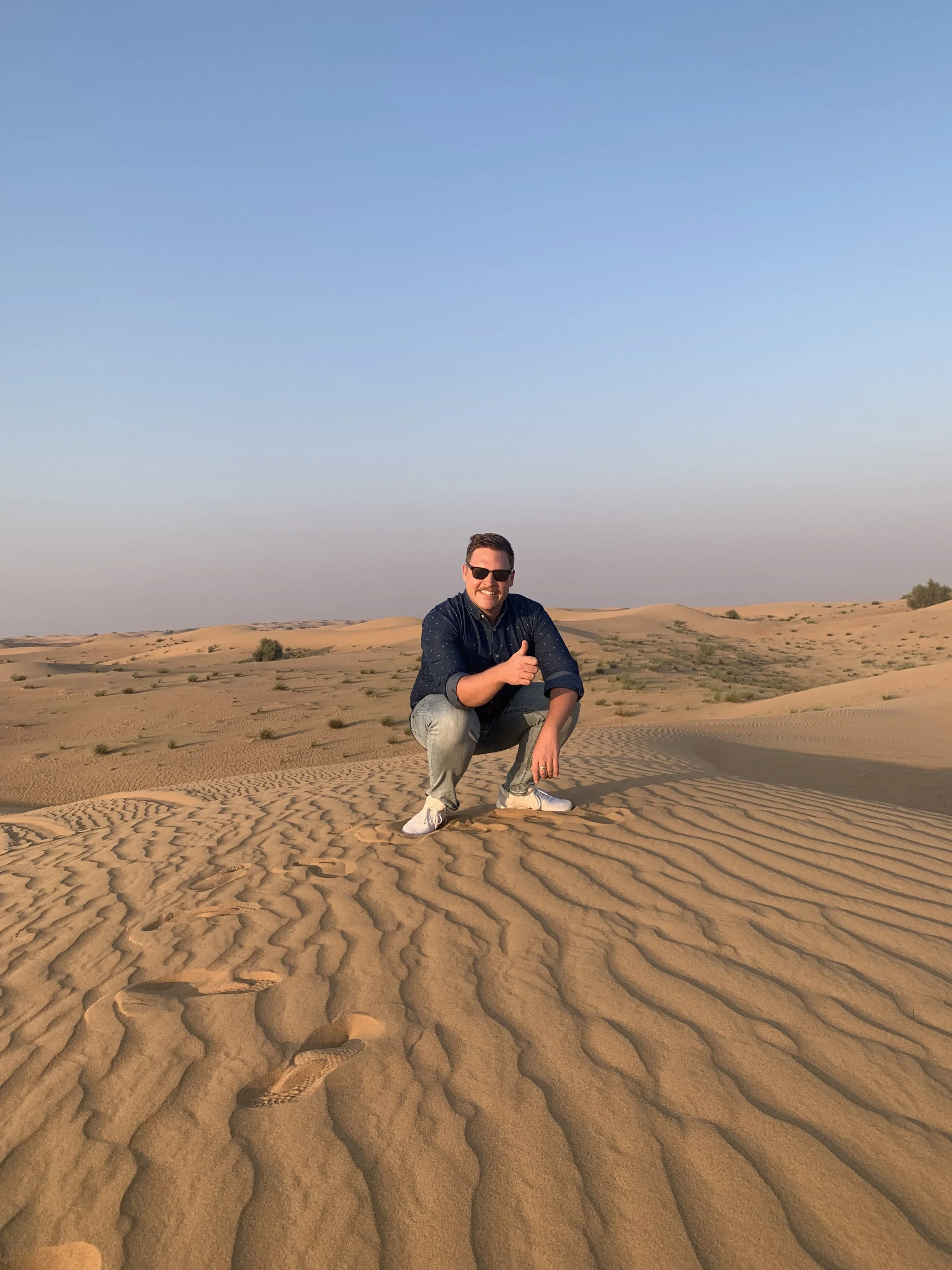 A man crouching in a desert with rippled sand dunes, wearing sunglasses, a dark shirt, light jeans, and white sneakers, giving a thumbs-up.