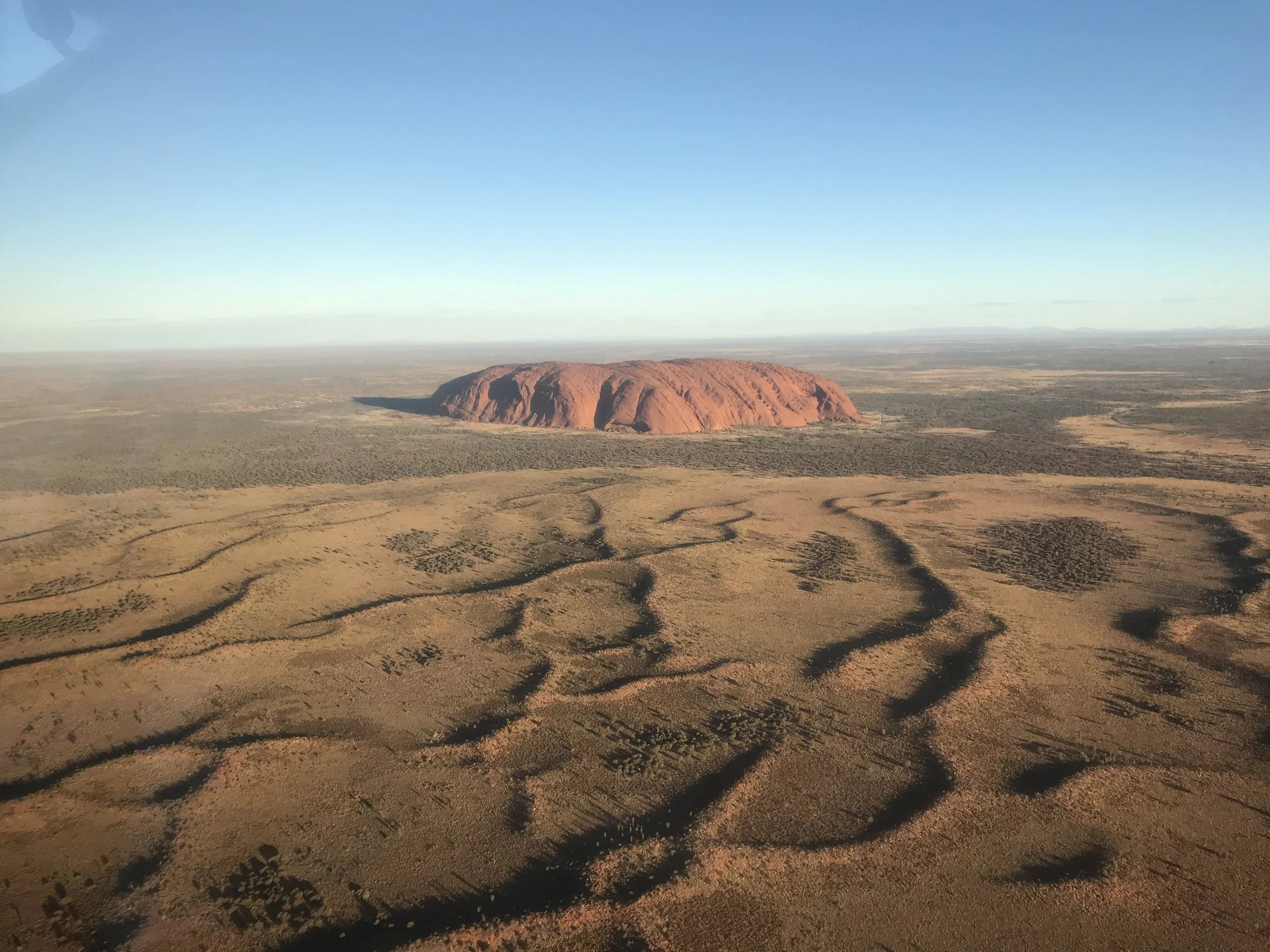 Red rock formation rising from a desert plain under clear sky, tyre tracks in the foreground.