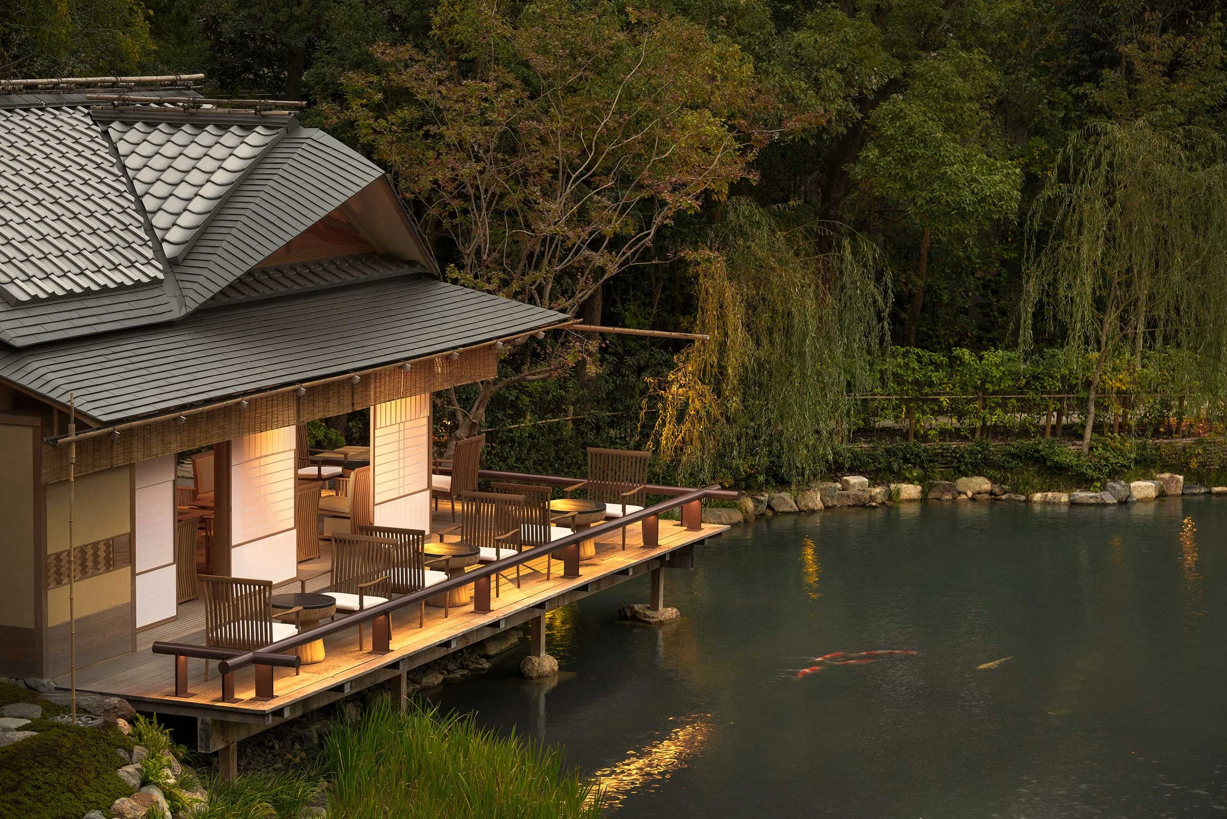 A traditional Japanese-style building with a tiled roof and a wooden deck overlooking a body of water, surrounded by lush green trees and bushes.