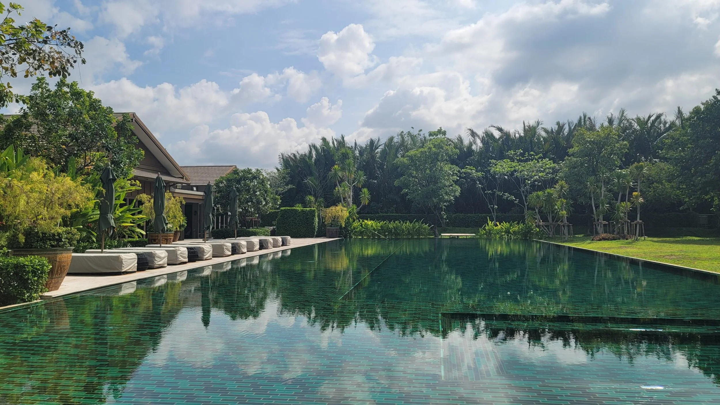 A luxurious outdoor swimming pool surrounded by lush greenery, with lounge chairs and umbrellas along the poolside, and a partly cloudy sky overhead.