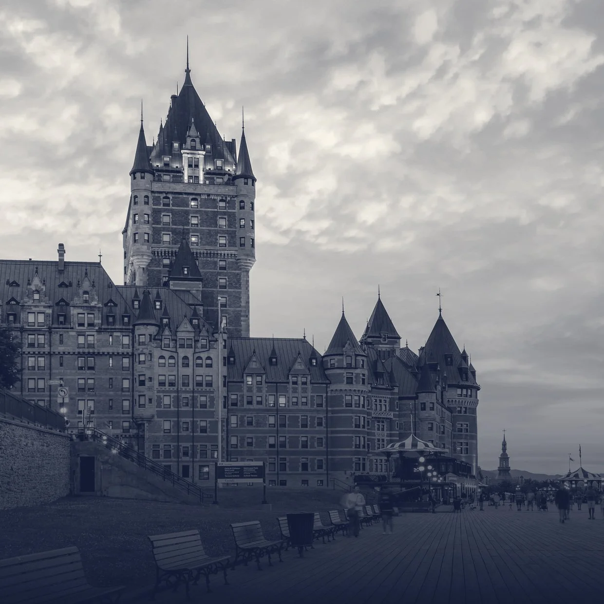 Historic castle with multiple towers and turrets, situated along a boardwalk with benches, under a cloudy sky, in a monochrome style.