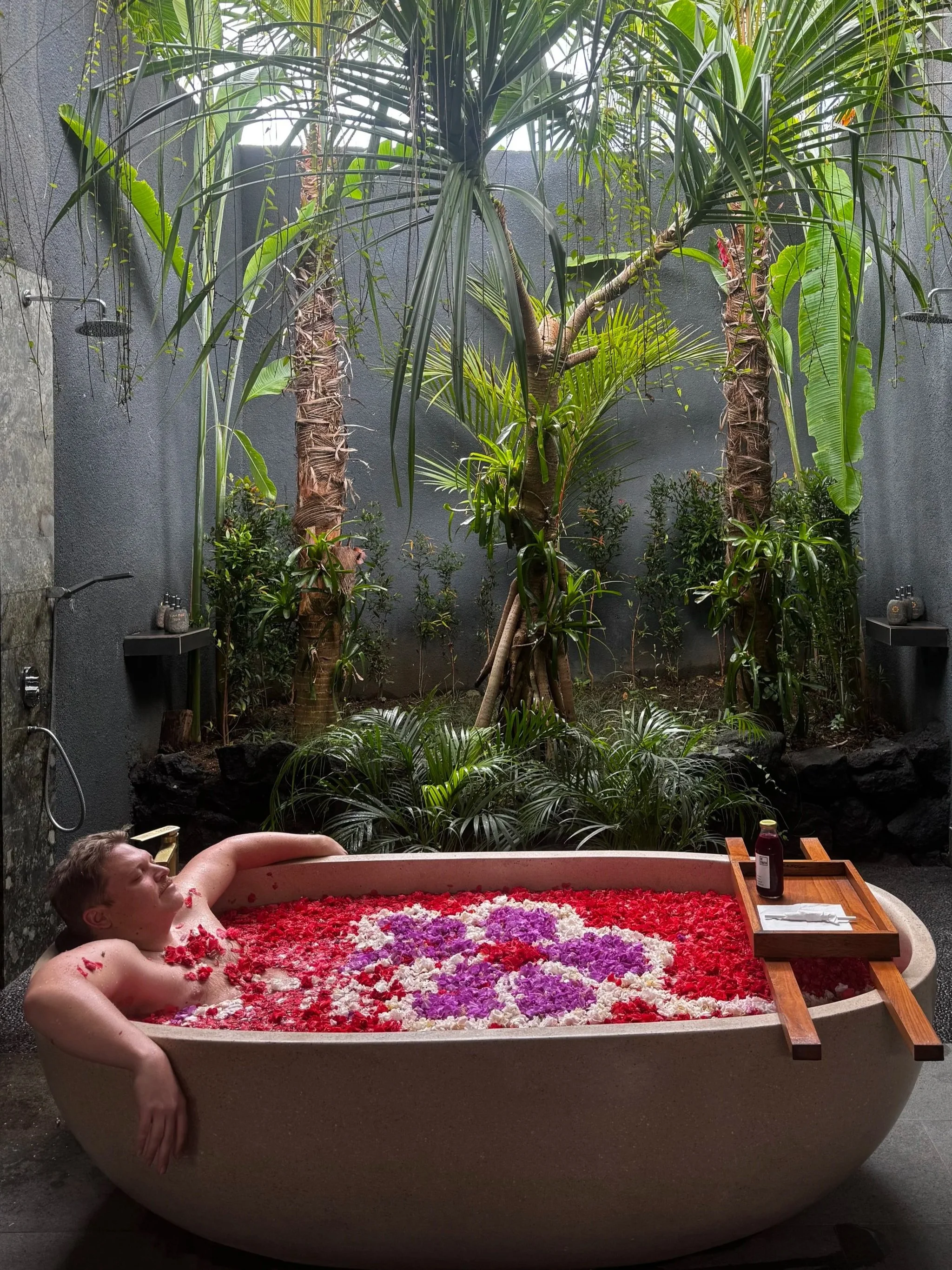 A young boy is relaxing in a bathtub filled with red, white, and purple flower petals, surrounded by a lush indoor garden with tropical plants and a gray wall.
