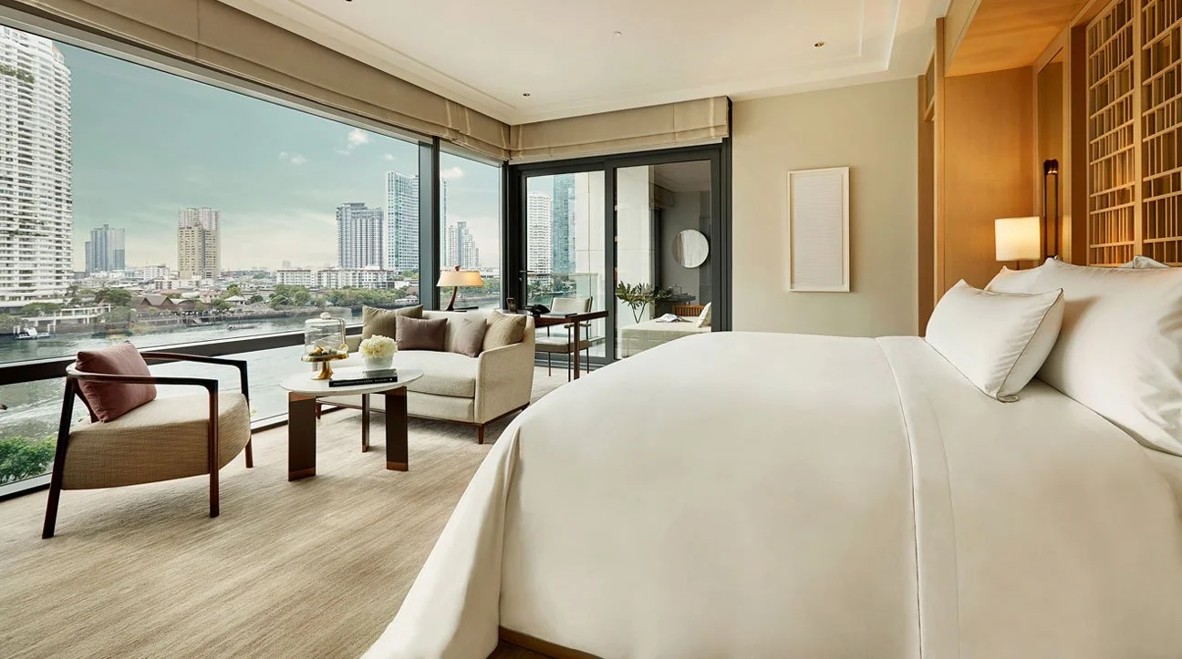 A hotel room with a large bed, white bedding, and a wooden headboard. There is a sitting area with a sofa, armchair, and coffee table near large windows that reveal a city skyline with tall buildings and a river.