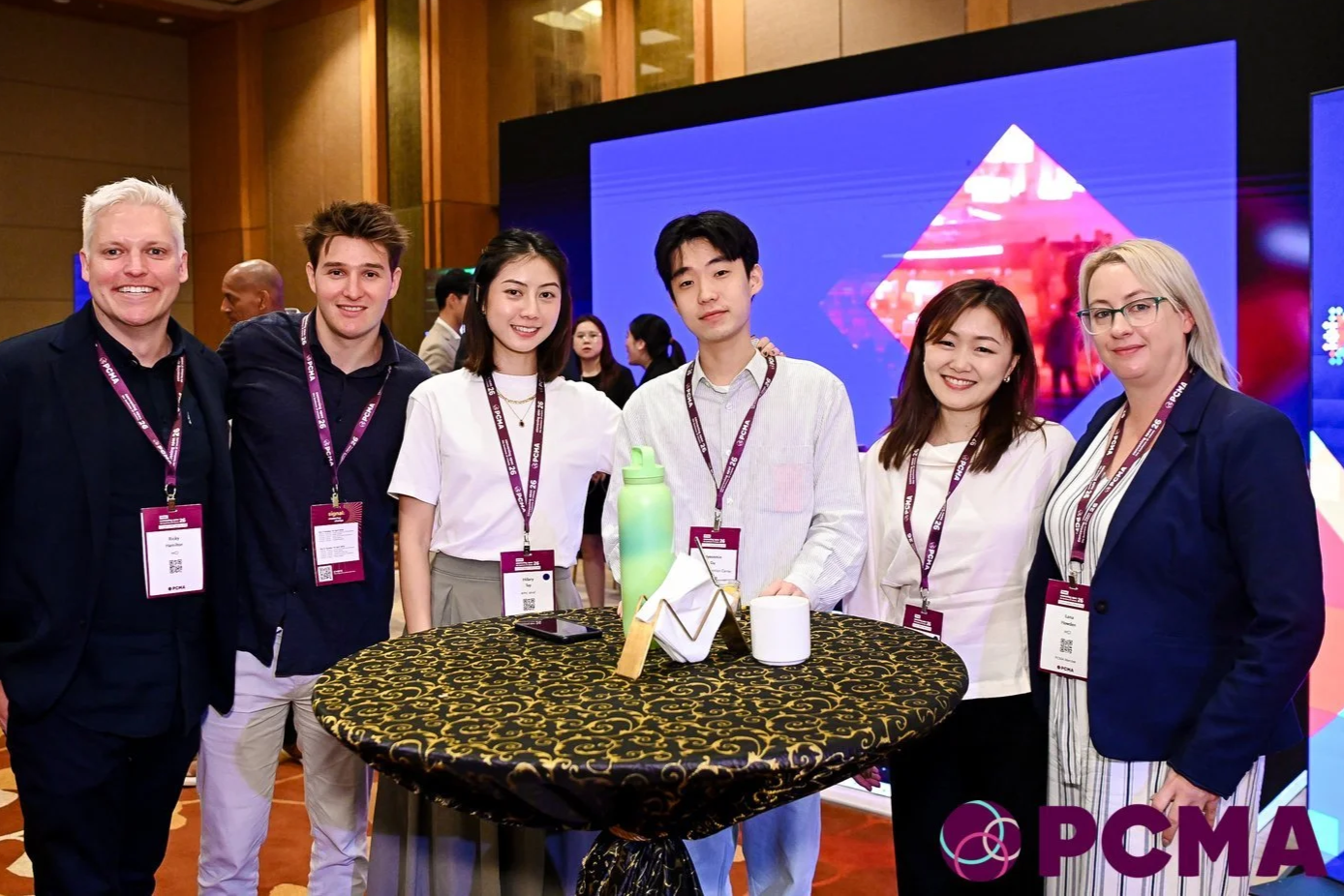 Six people standing together at a conference, smiling and posing for a photo behind a round table with a black and gold tablecloth. They are wearing conference name tags. There is a large digital screen in the background with colorful graphics.