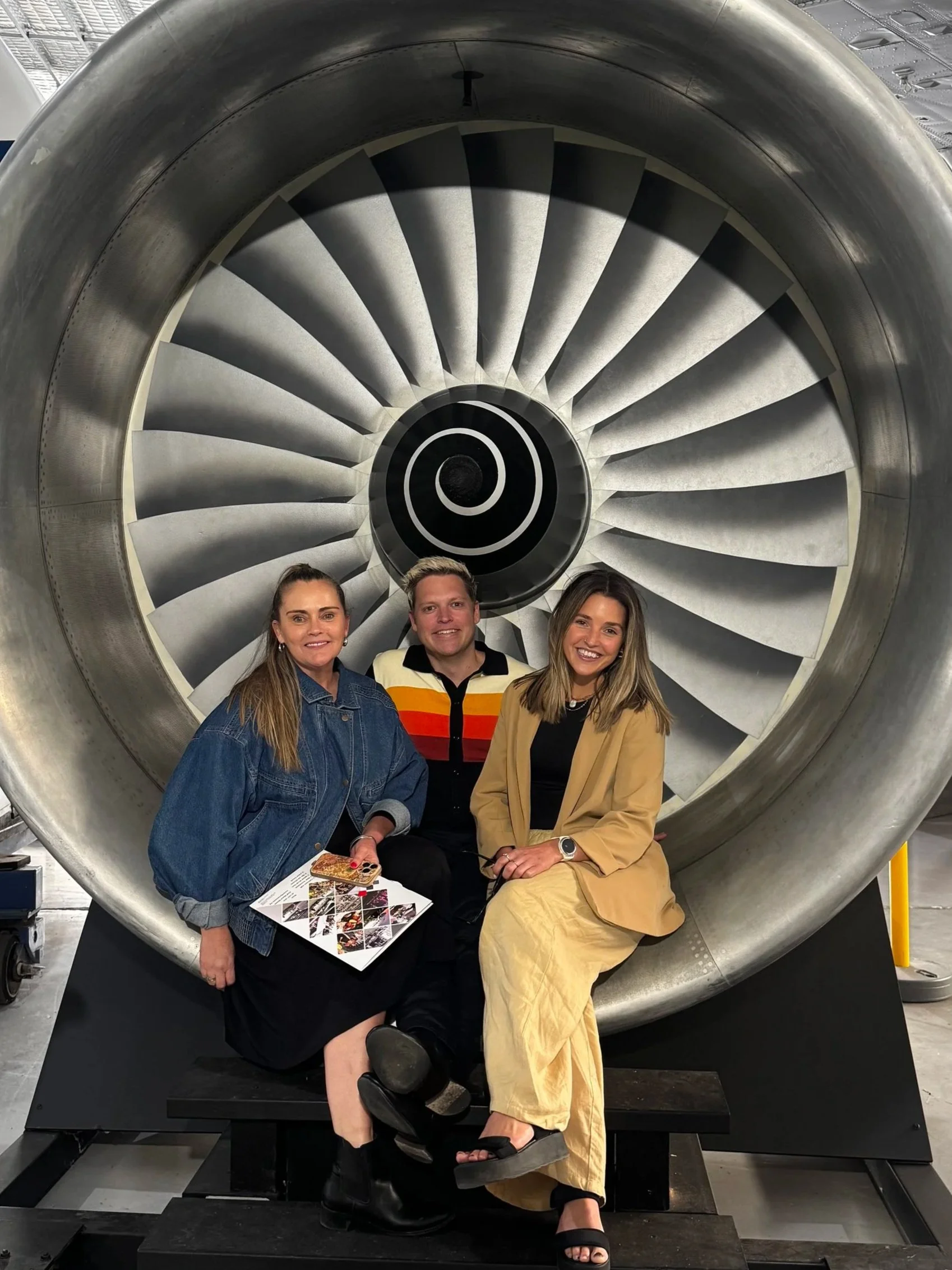 Three people sitting inside a large jet engine display, with a spiral pattern visible at the center. The two women are smiling, and the man in the middle is also smiling, holding a brochure or pamphlet. The woman on the left is wearing a denim jacket, the man is wearing a colorful striped shirt, and the woman on the right is wearing a beige jacket.