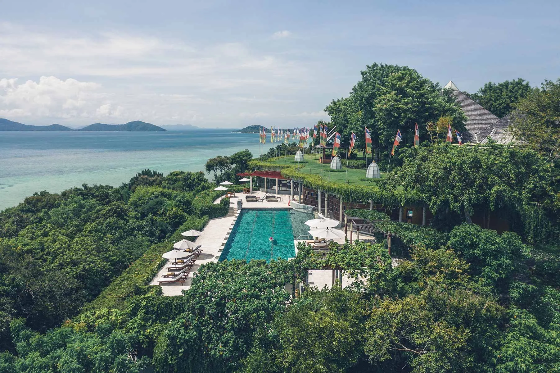Luxury hotel resort with a swimming pool, surrounded by greenery, overlooking the ocean and distant islands, with flags lined along a hillside
